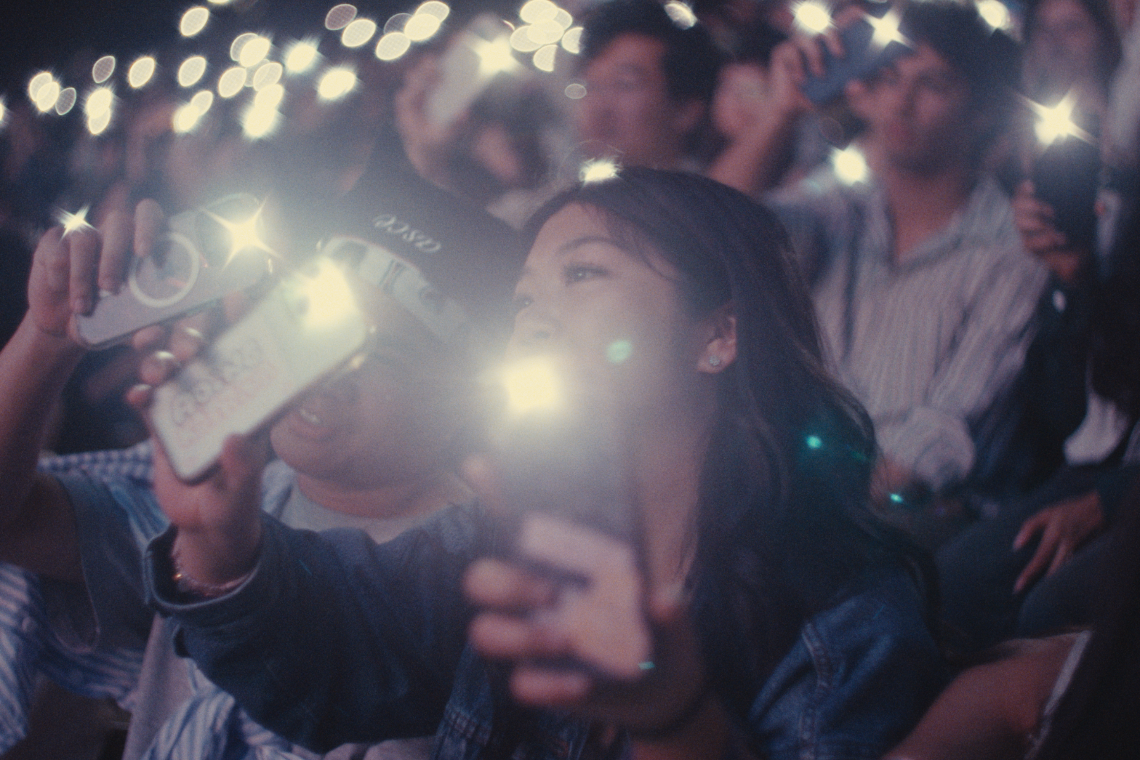 A group of people at a concert or event taking photos or videos with their phones and cameras, with lights and flashes creating a bokeh effect.