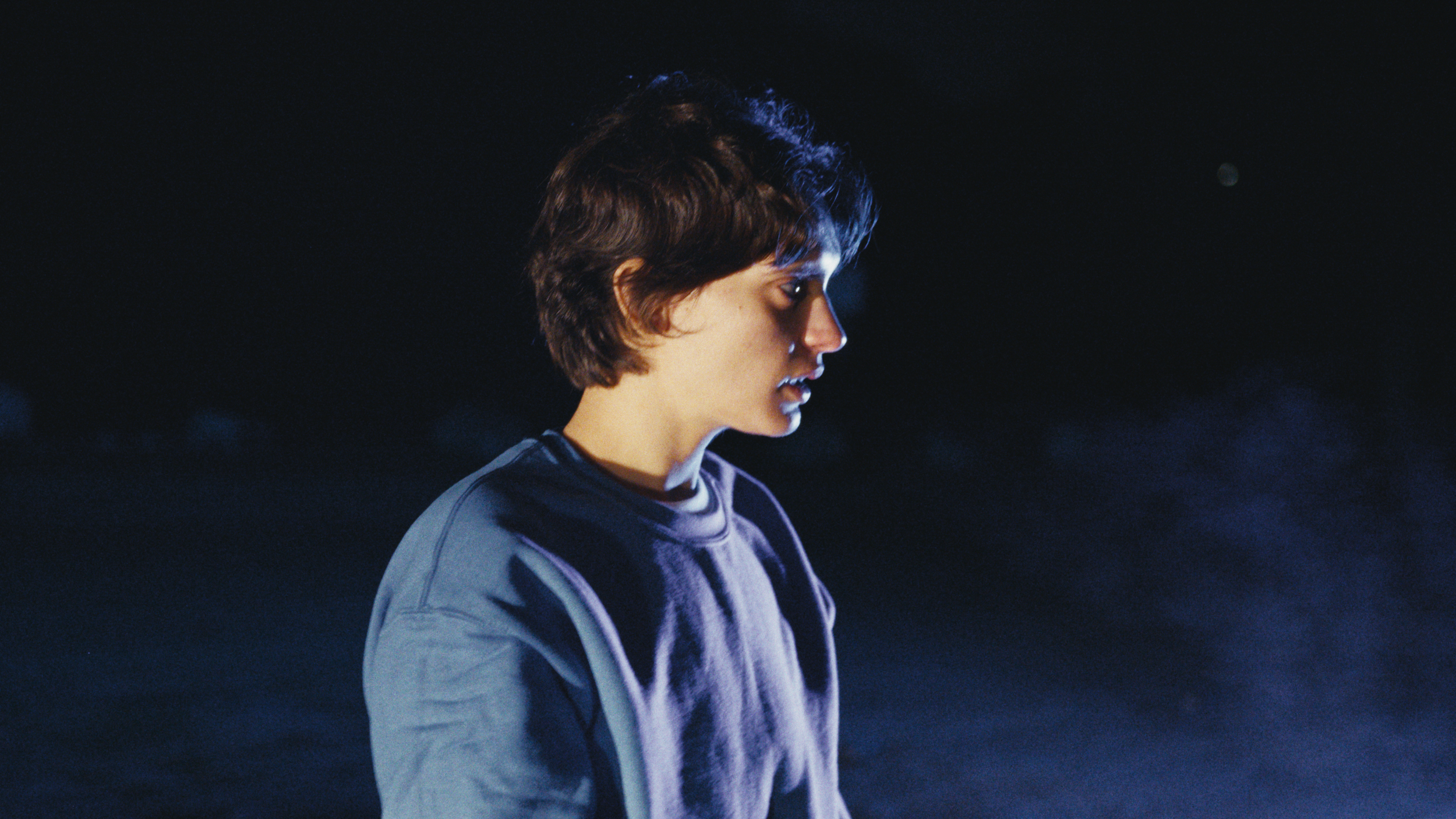 Side profile of a young person with short, dark hair, wearing a light jacket and dark shirt, looking downward against a dark, night sky background.