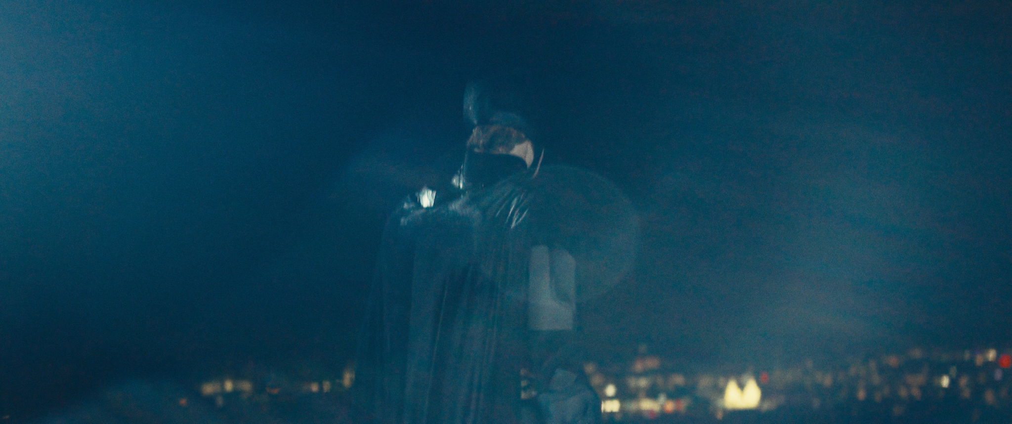 A person wearing a hoodie and a face mask, standing outdoors at night, with city lights blurred in the background.