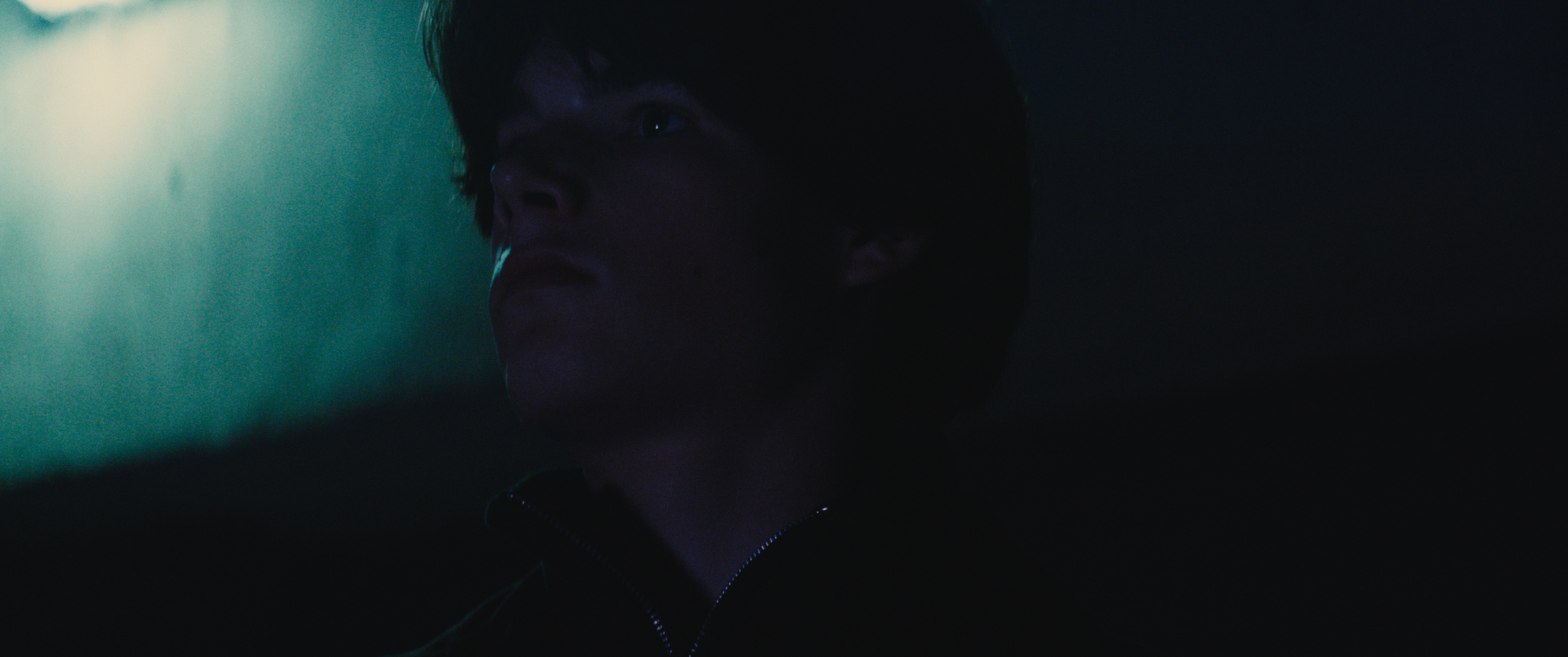 A young person with dark hair looking to the side in a dark environment, illuminated by subtle blue and green lighting.