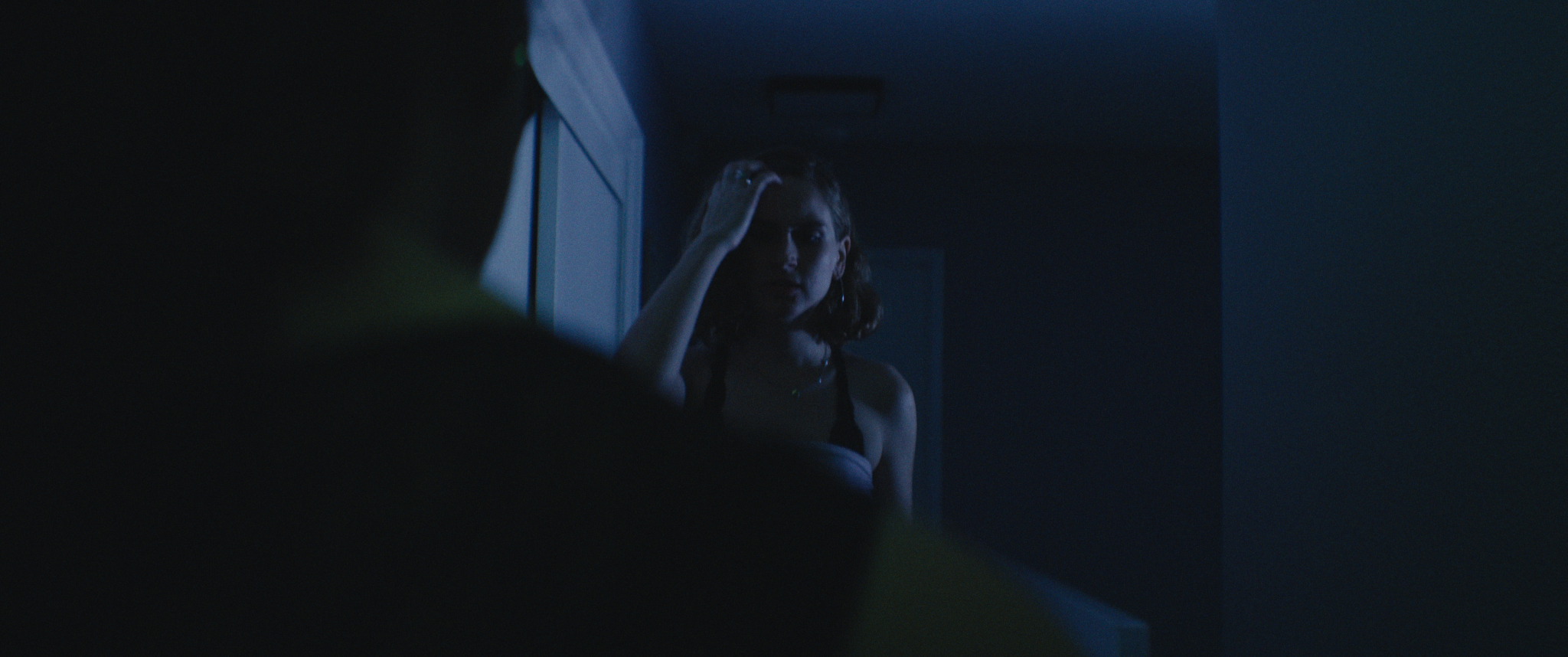 A woman with shoulder-length hair looking into a mirror, touching her forehead, in a dimly lit room with a blue tone.