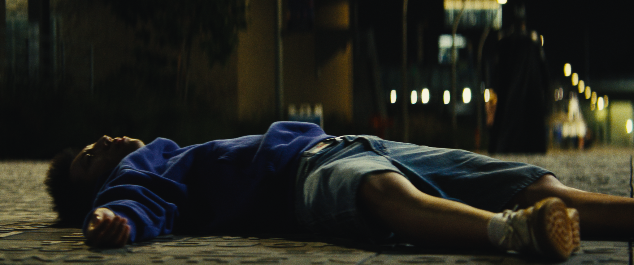 A young boy lies on the ground at night, wearing a blue jacket and shorts, with his eyes open, on a paved surface in a dimly lit area.