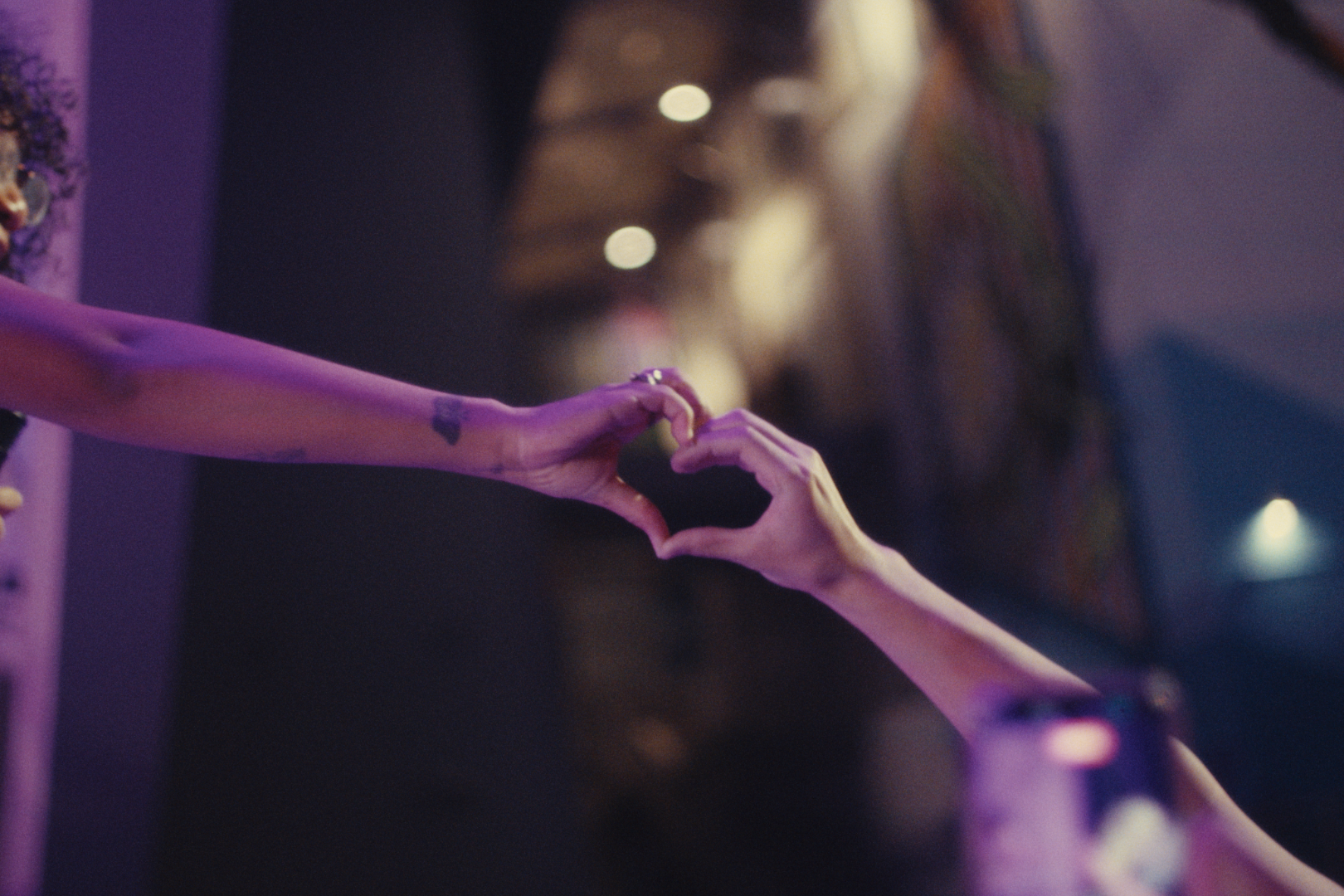 Two hands forming a heart shape against a dark, blurred background with purple lighting.