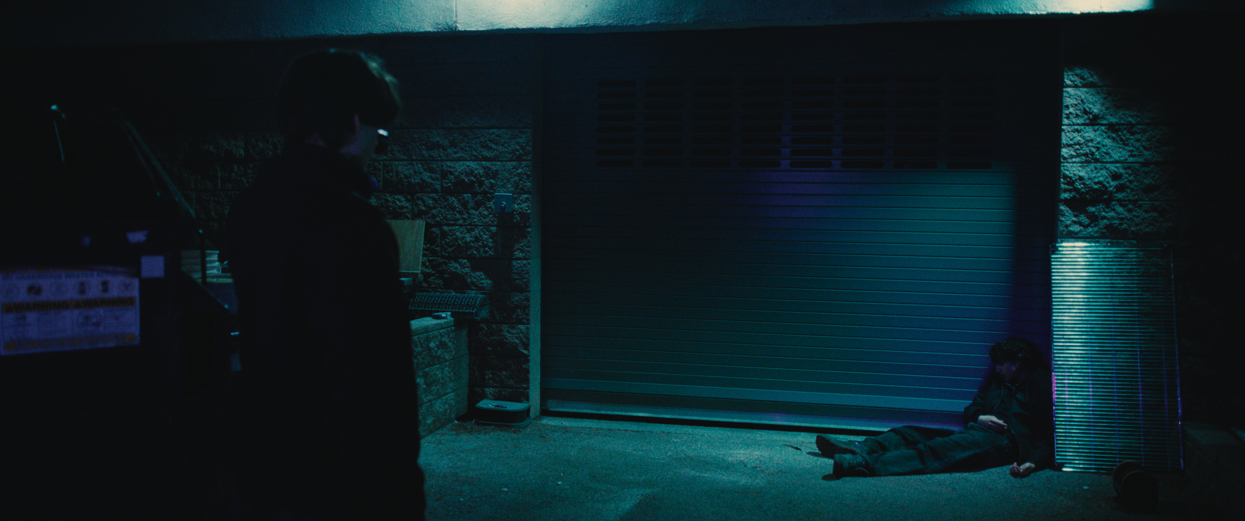 A person sitting on the ground with a phone at night, illuminated by blue and purple light, with a closed metal shutter behind them. Another person stands nearby, partially visible, in a dark environment.
