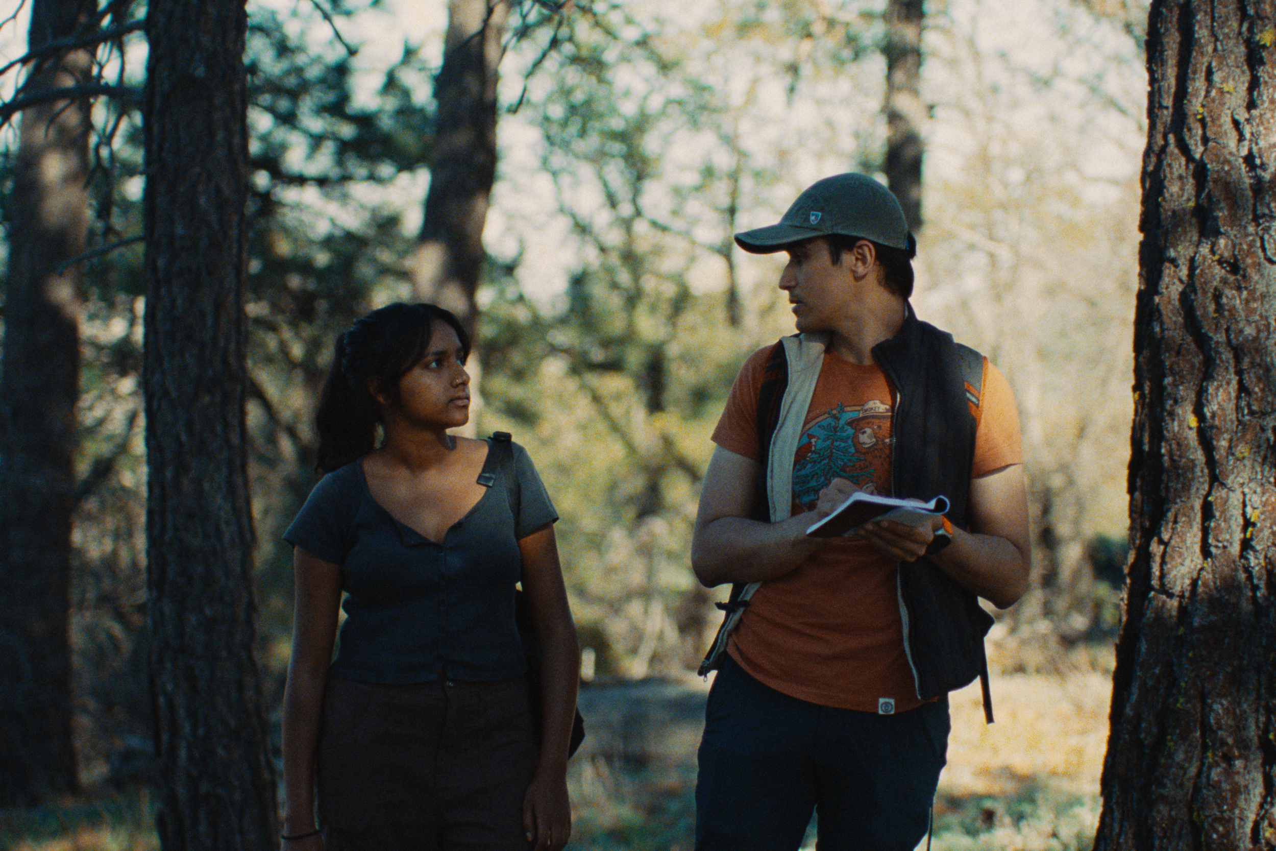 Two young people, a woman and a man, walking in a forested area, engaged in a conversation. The woman has dark hair and is wearing a dark shirt, while the man is wearing a cap and a backpack, holding a notebook or map.