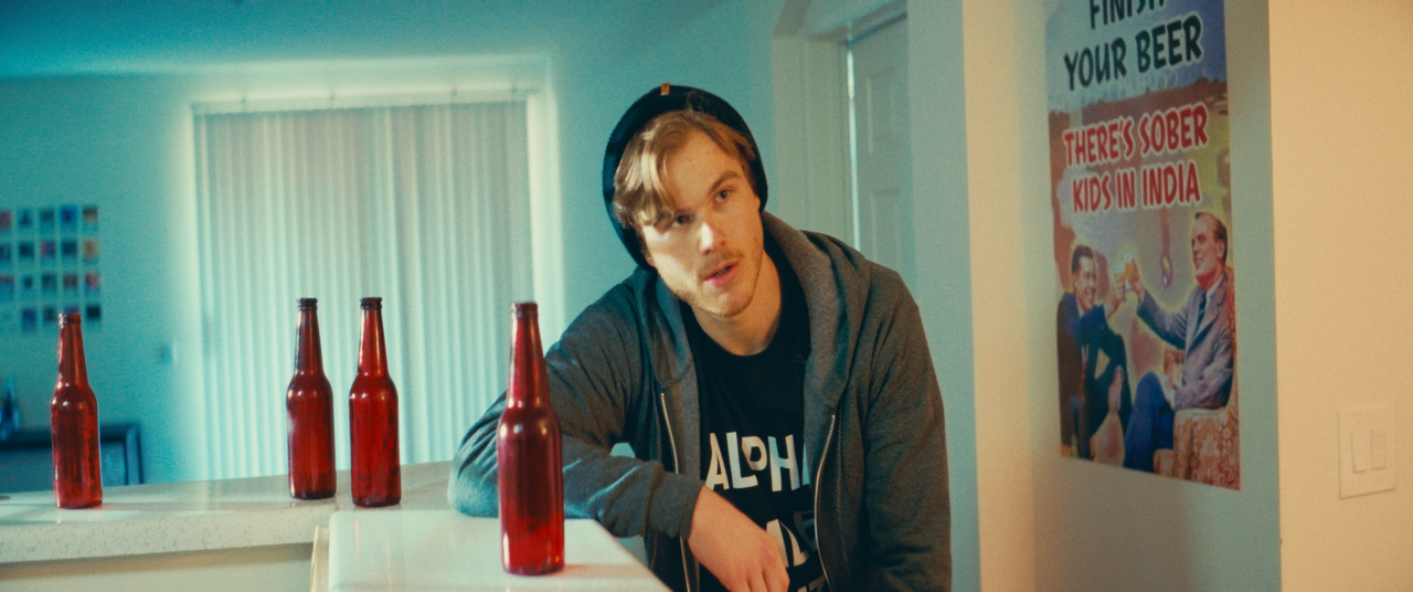 A young man with light hair and a mustache wearing a gray hoodie and black beanie, leaning on a counter with four red bottles, in a room with a poster on the wall that reads 'Finish Your Beer, There's Sober Kids in India.'