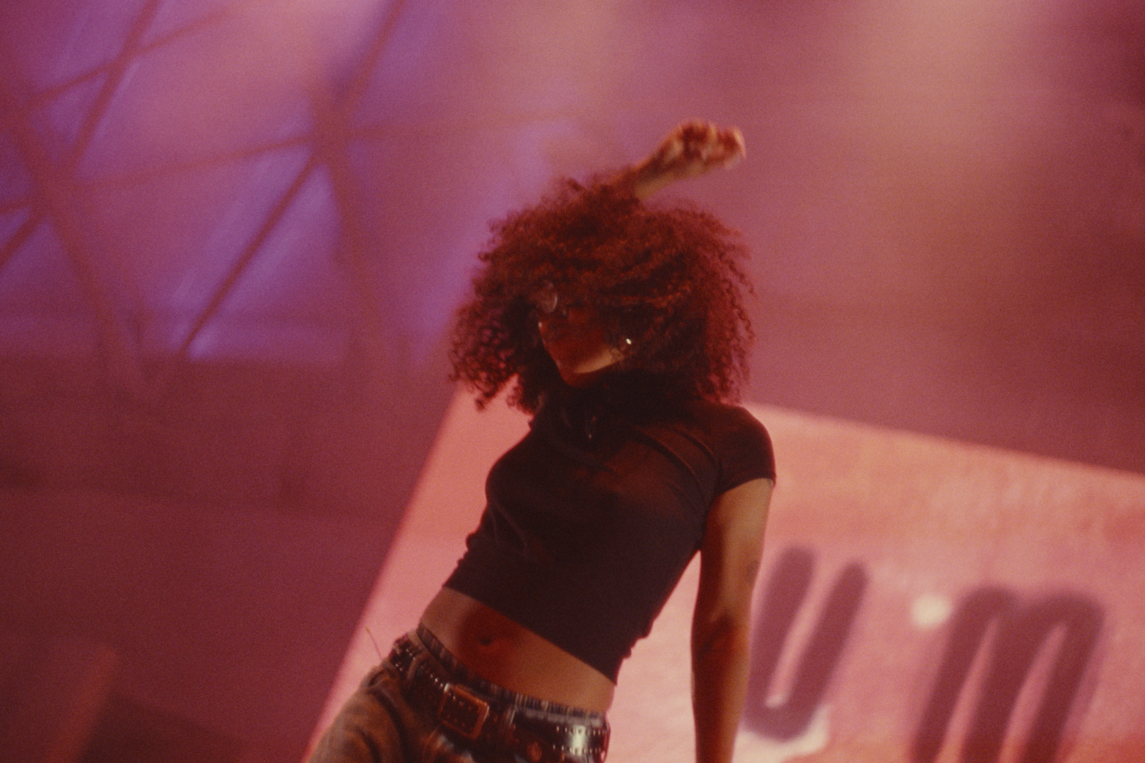 A young woman with curly hair dancing in a dimly lit area with pink and purple lighting.