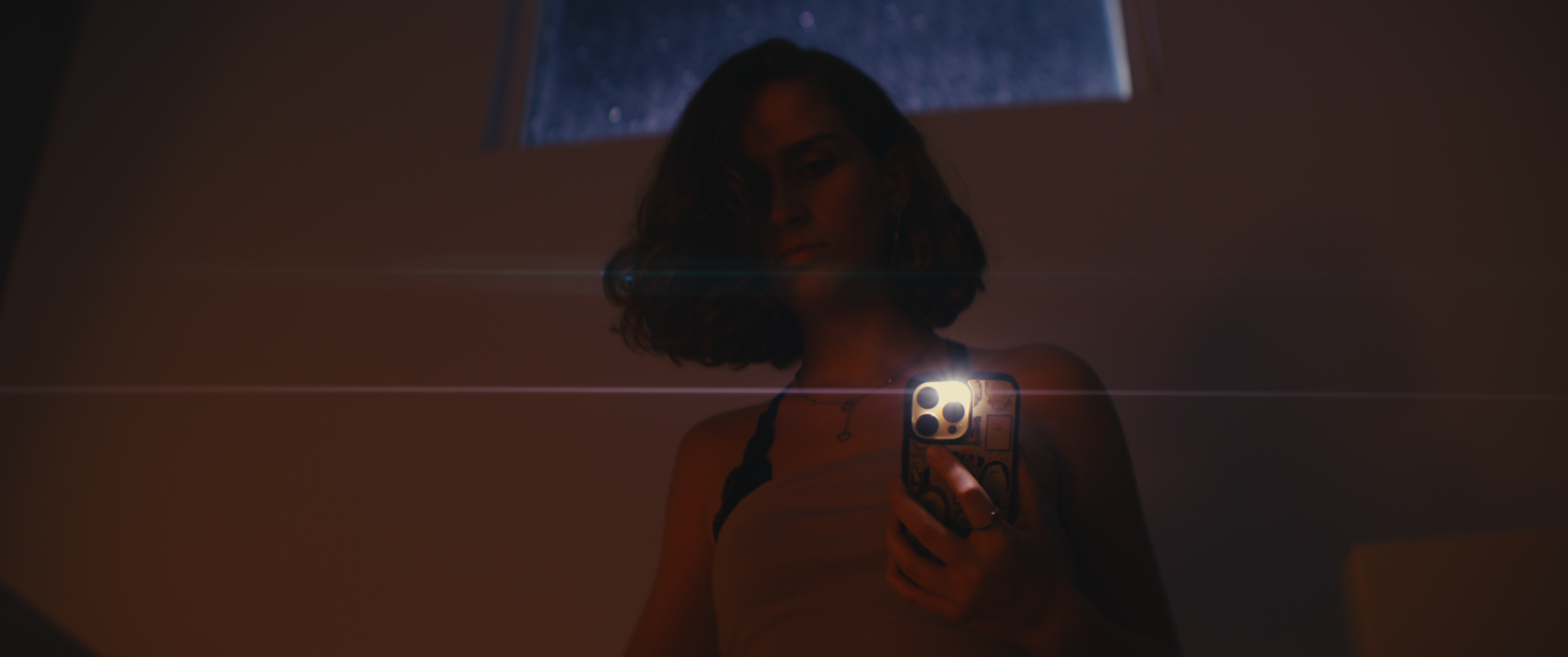 A woman taking a mirror selfie with a smartphone in a dimly lit room, with a window in the background showing a starry sky.