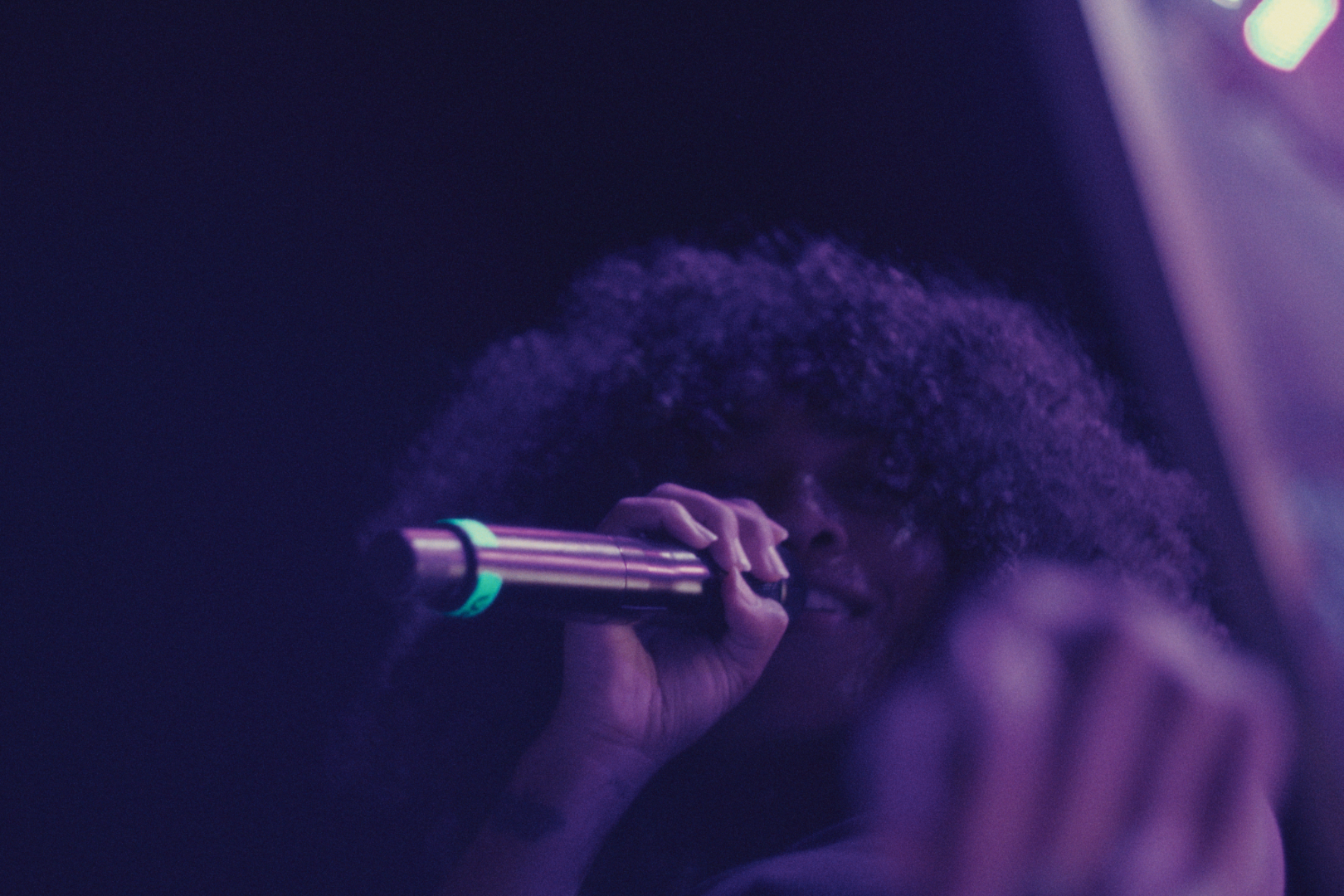 A woman with large curly hair looking through a telescope, illuminated by violet and purple lighting.