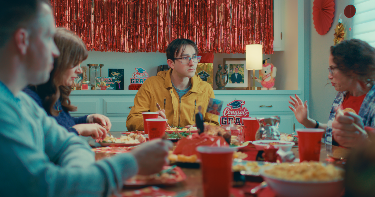 Group of people sitting around a decorated table with food, celebrating a graduation at a party with festive decorations, including a 'Congrats Grad' sign, and party supplies.