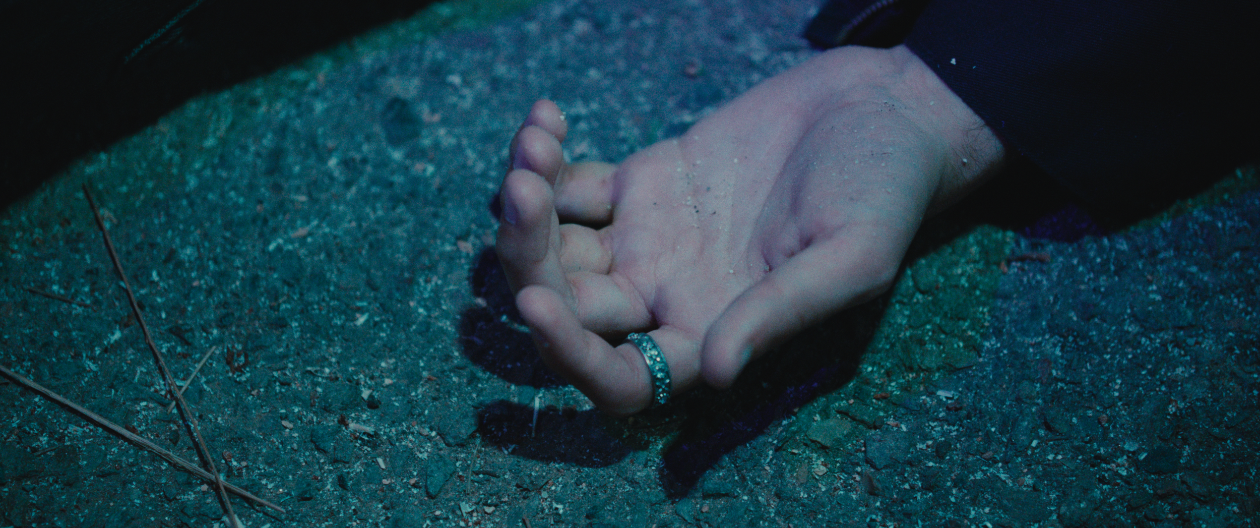 Close-up of a hand with a silver ring, lying on gravel ground, with a dark sleeve visible.