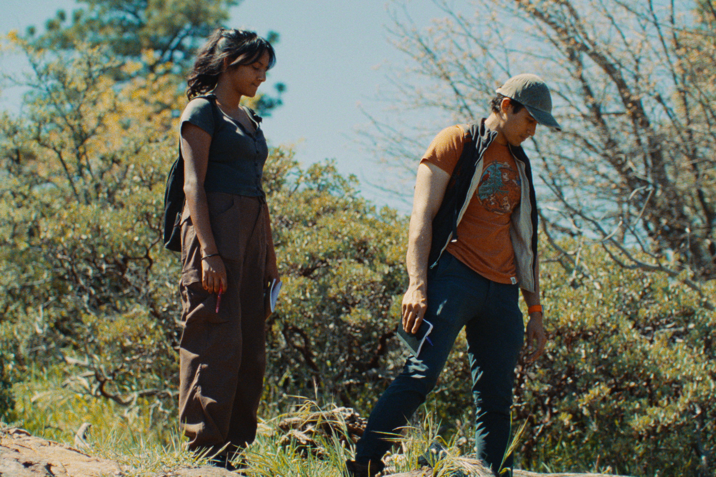 Two young people, a woman and a man, walking outdoors on a sunny day amidst trees and bushes, with the woman holding some papers.