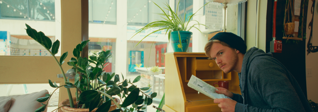 A young man wearing a black beanie and gray hoodie sitting at a counter, looking at a newspaper, with potted plants and a window behind him.