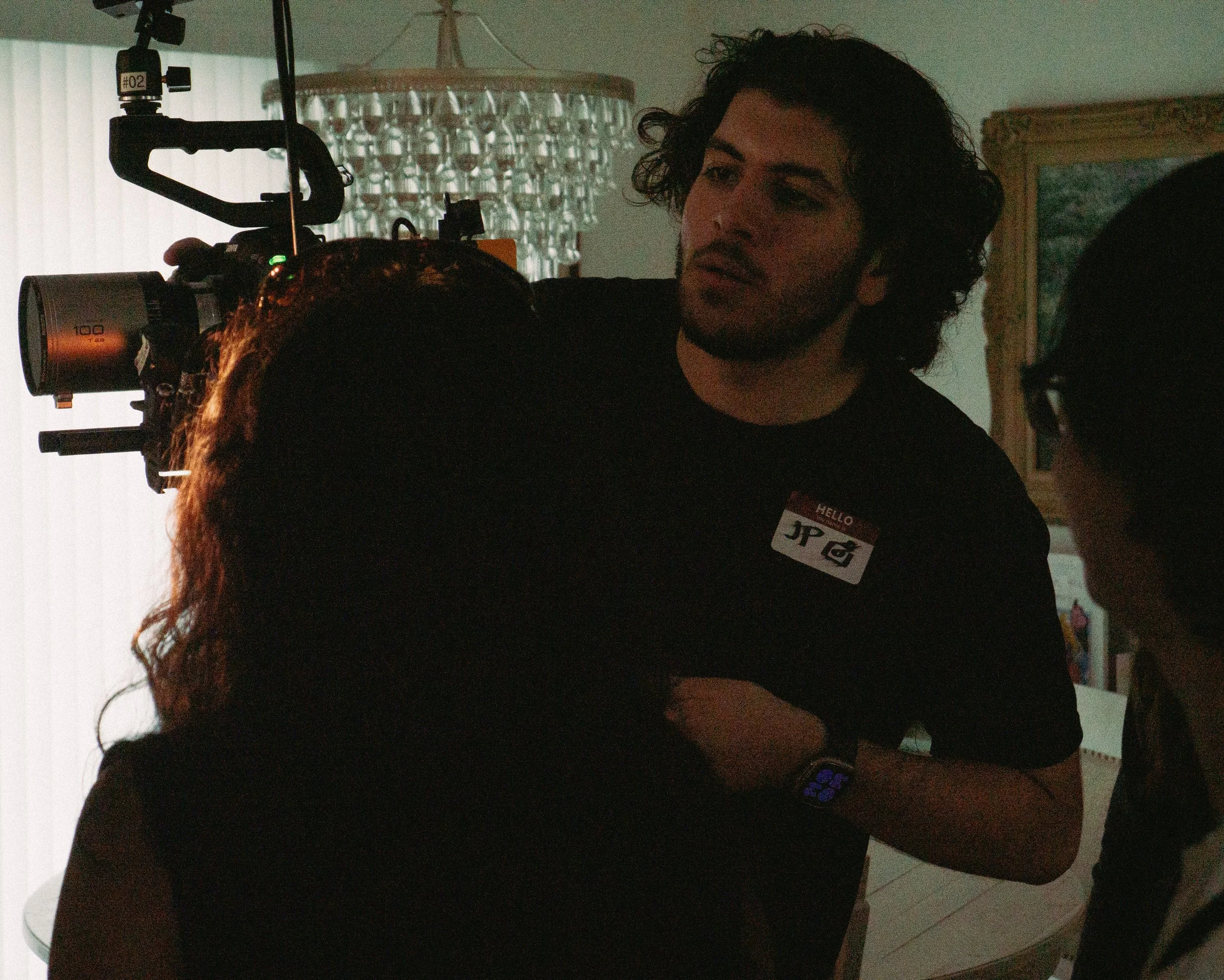 A man with curly hair and a beard wearing a black shirt, holding a camera with a large lens, standing inside a room. There is a name tag on his shirt with the name 'JP'. A chandelier hangs from the ceiling in the background, along with window blinds and a framed picture.