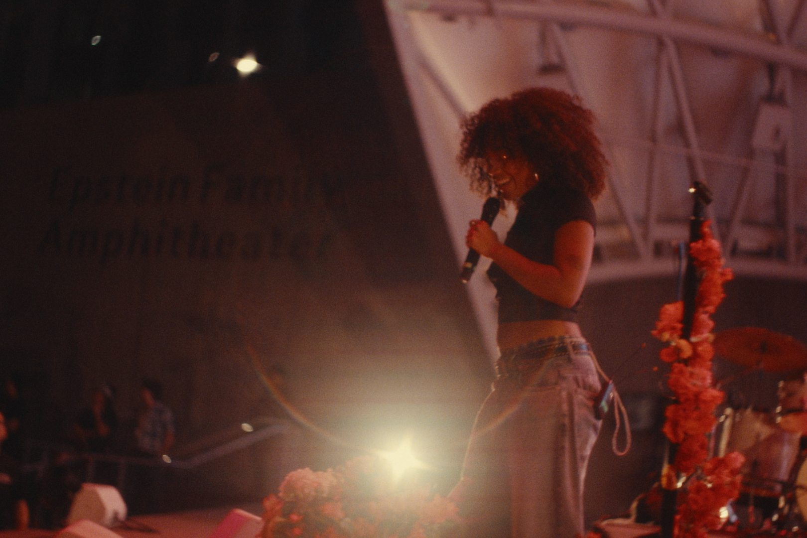 A woman with curly hair and glasses holding a microphone on a stage, smiling, dressed in a black crop top and jeans, with a tropical-themed microphone stand decorated with pink flowers.