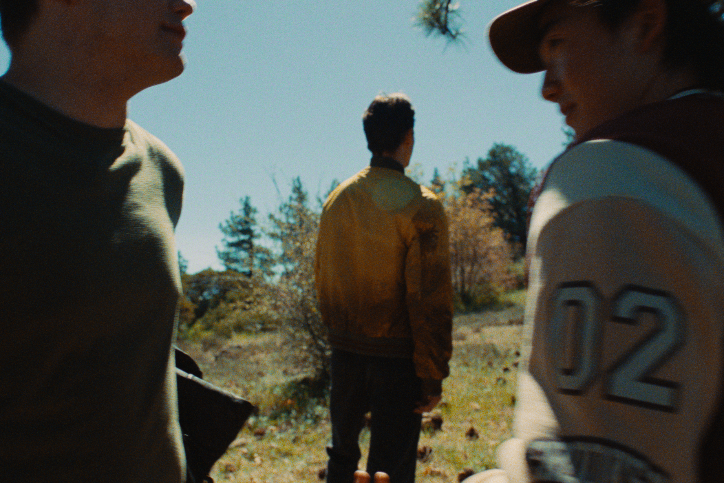 Three people standing outdoors, partially visible, with two in the foreground and one in the background, under a clear blue sky, with trees and foliage around.