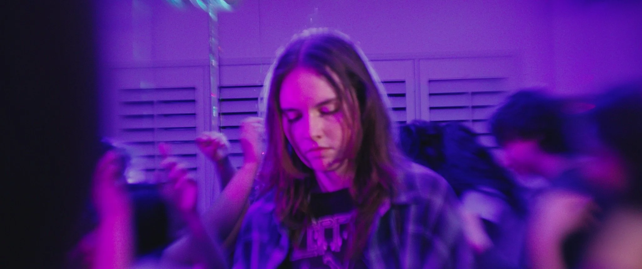 A young woman with shoulder-length hair, wearing a plaid shirt, standing in a room illuminated with purple and blue lights at a social gathering or party.