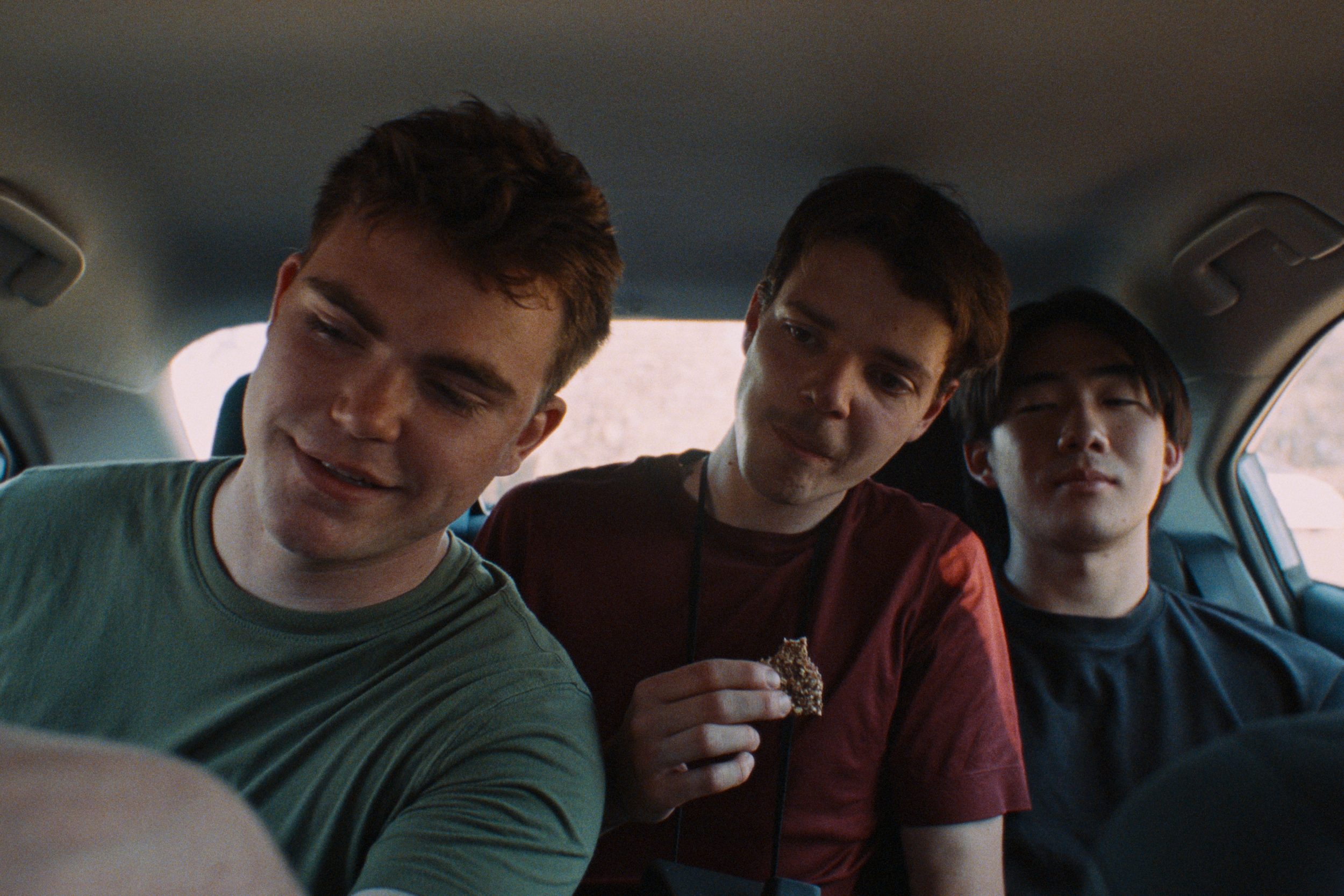 Three young men sitting in the back of a car, one is smiling and looking down, another is looking at him with a neutral expression, the third is focused on something outside the frame, one is holding a rock. All three are casually dressed.