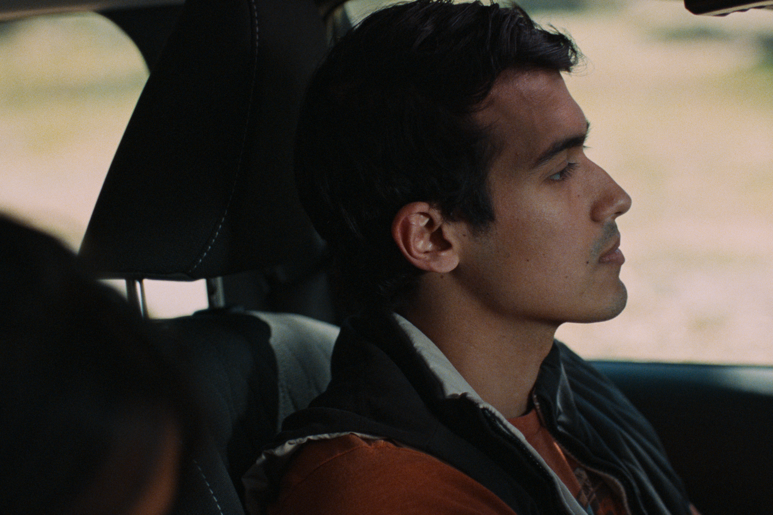 A person with short dark hair sitting in the driver's seat of a vehicle, looking ahead with eyes closed.
