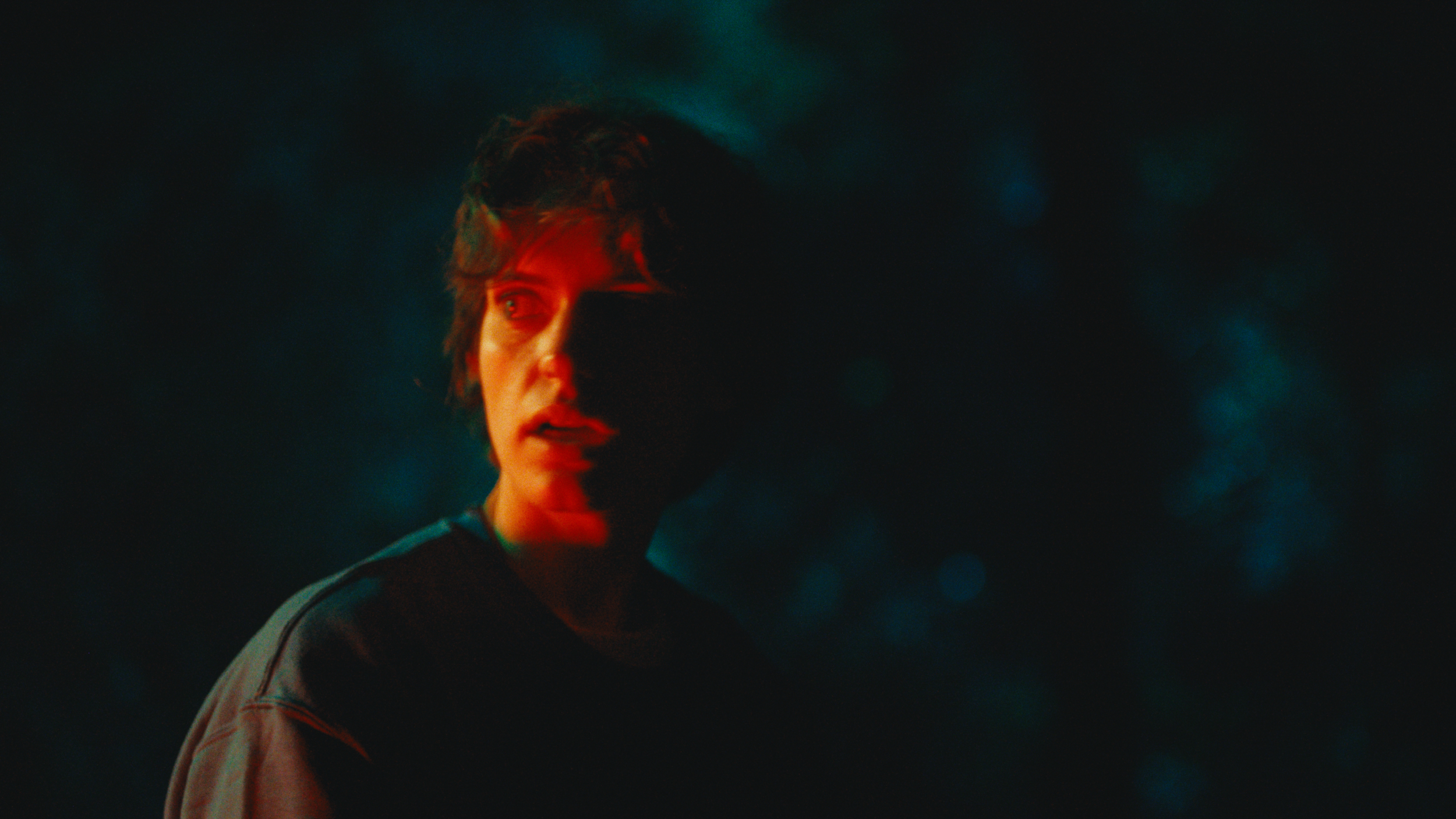A woman with short hair looks to the side in a dark, moody setting with colored lighting creating a shadowed effect on her face.