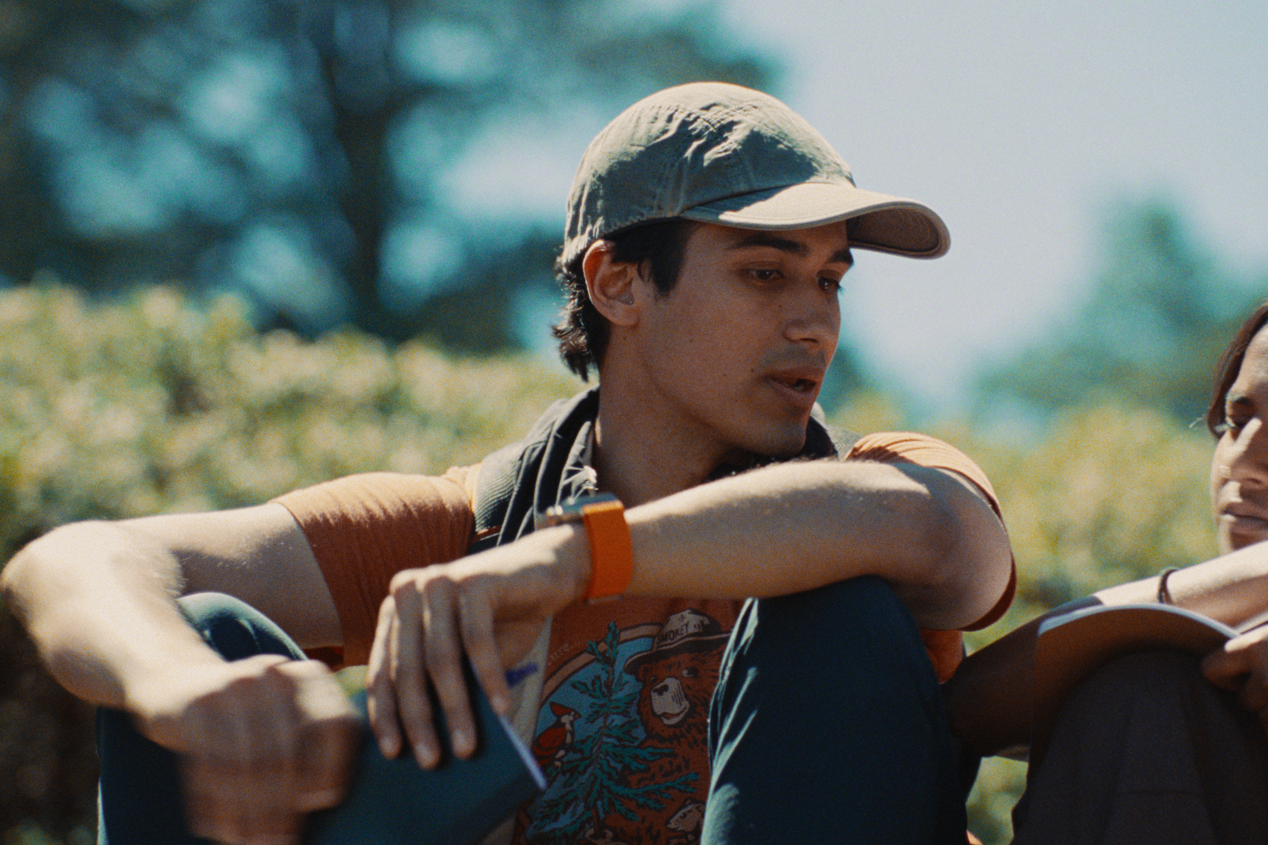 A young man wearing a beige cap and a colorful shirt, outdoors with blurred trees in the background.