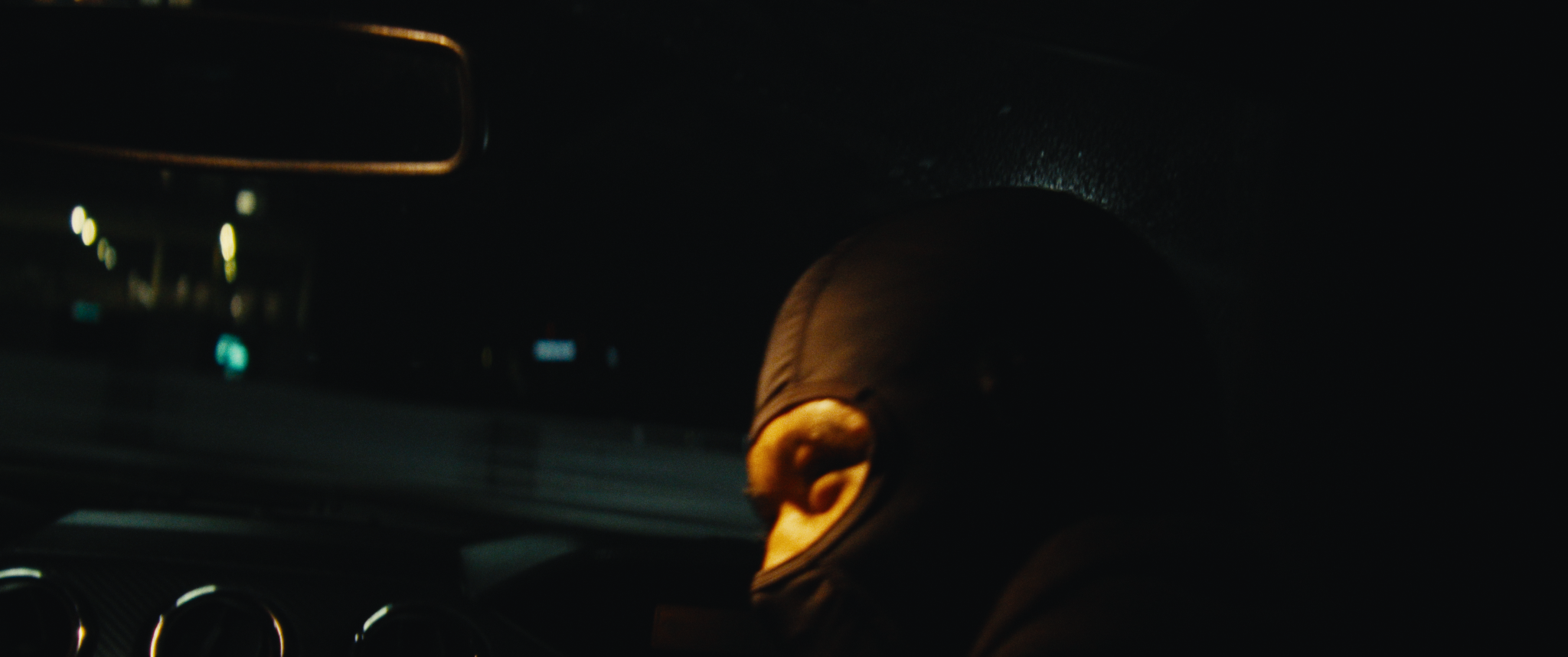 Close-up of a person's ear and mannequin head in a dark car interior, with a dashboard and illuminated controls faintly visible.