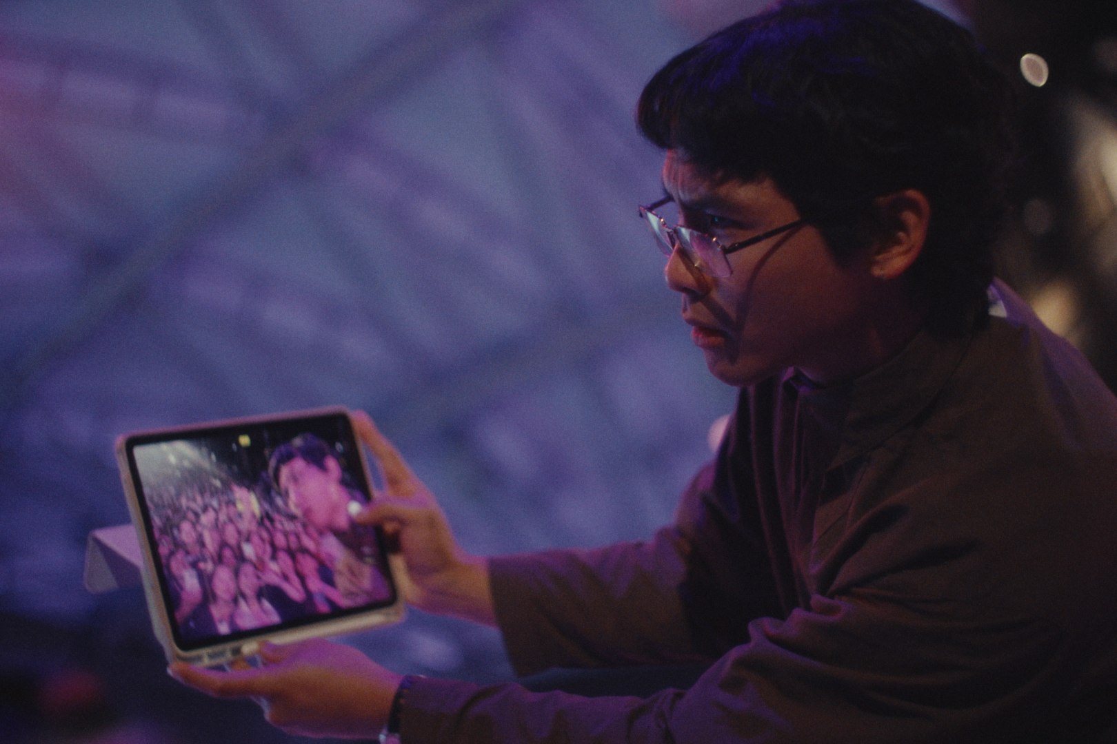 A man with glasses takes a selfie with a tablet in a dimly lit indoor space with purple and pink lighting.