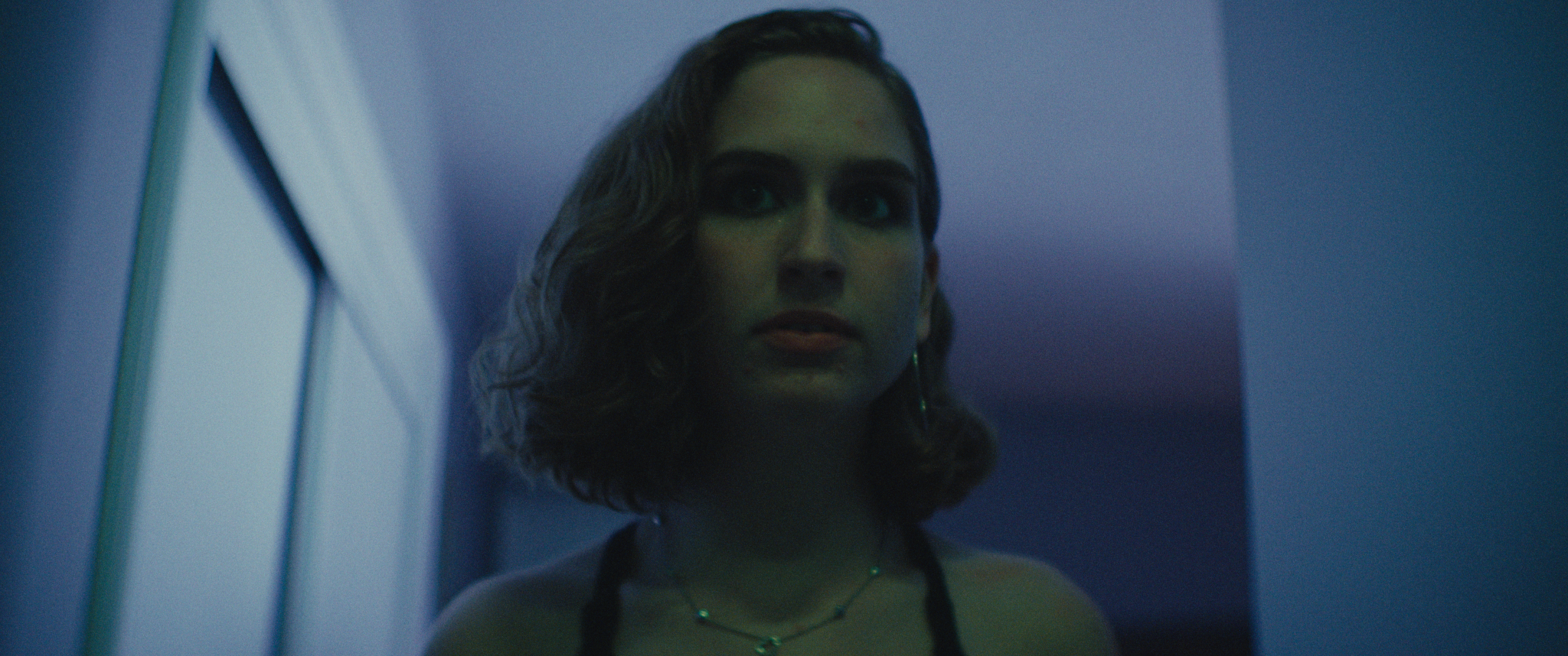 A woman with wavy hair and greenish skin standing in dim blue lighting, looking to the side.