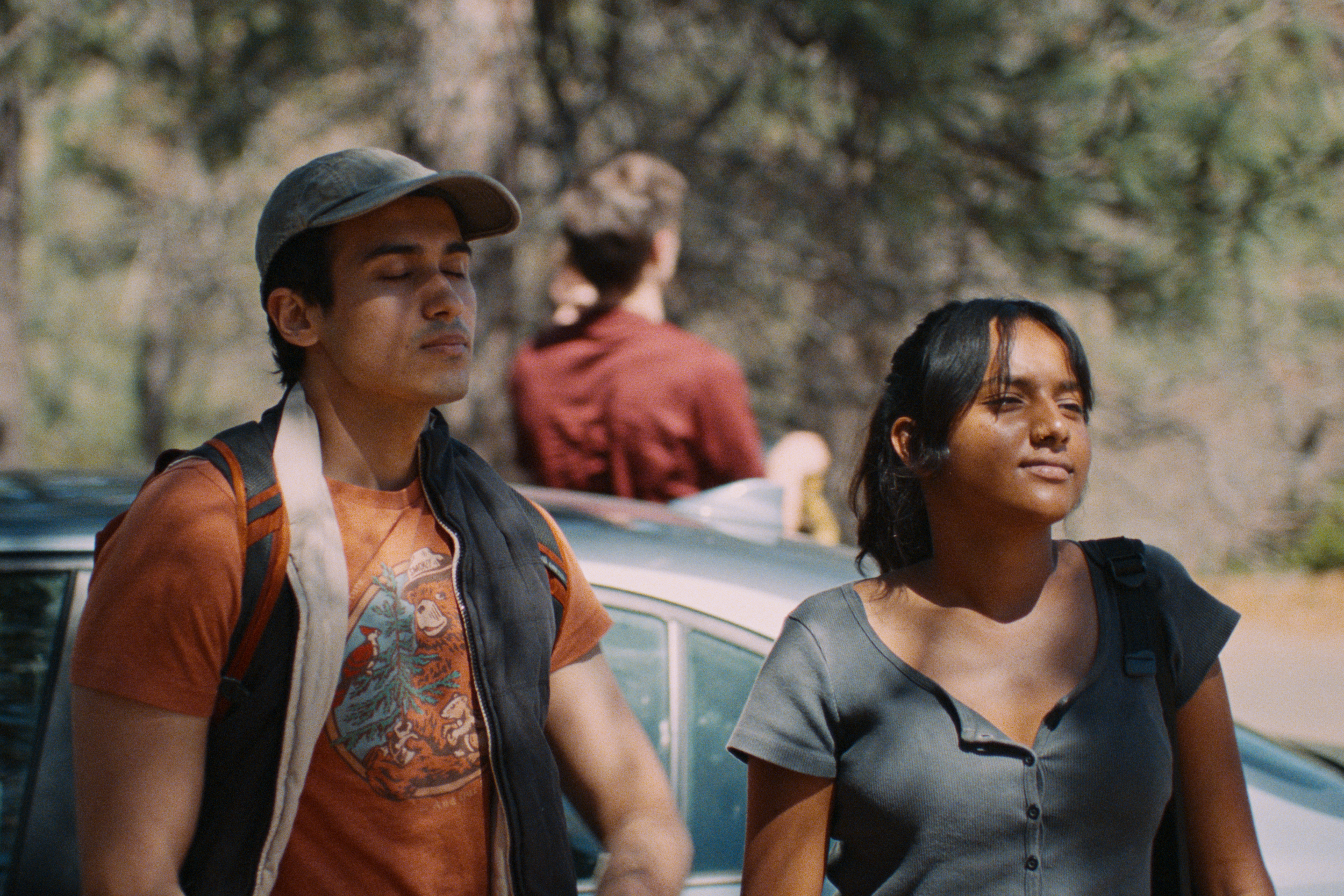 Two young women with backpacks walking outdoors, one with eyes closed, in front of a tree and a vintage car.