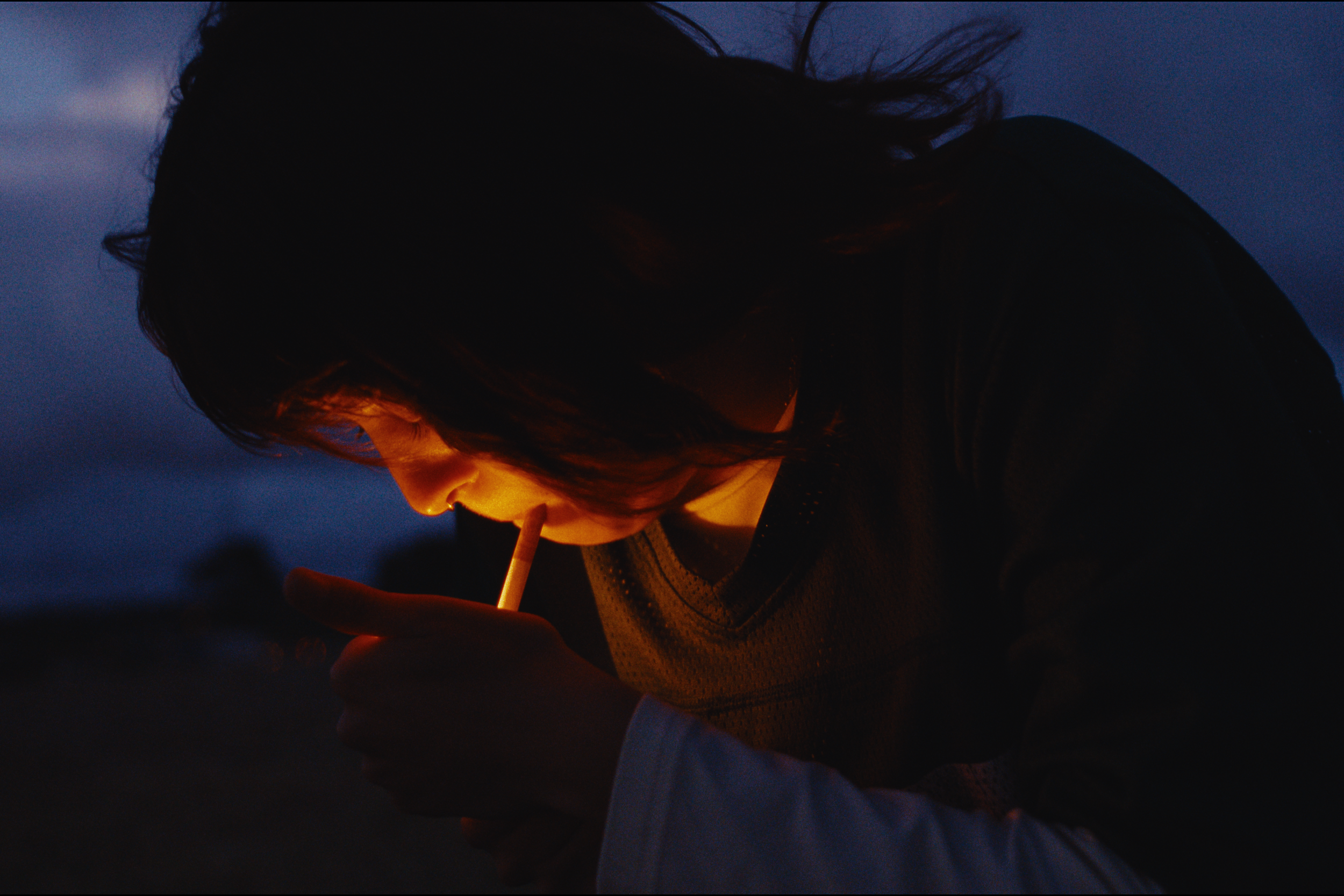 A person with shoulder-length dark hair smoking a cigarette at dusk or night, with a dark sky in the background.