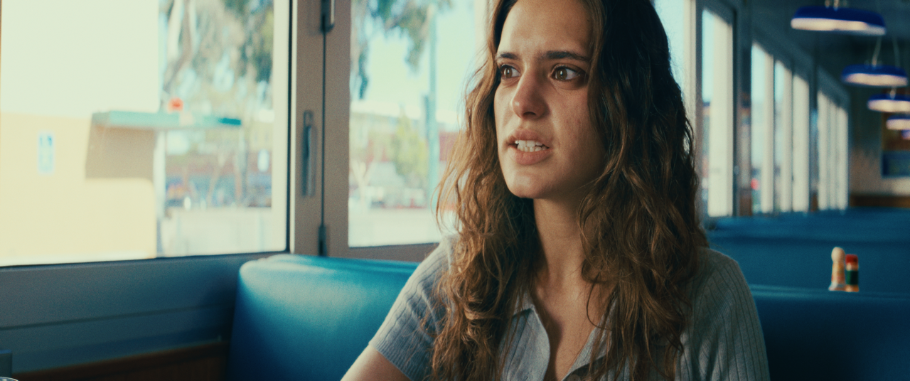 A woman with long, curly brown hair sits in a retro diner, looking out the window with a thoughtful or concerned expression.