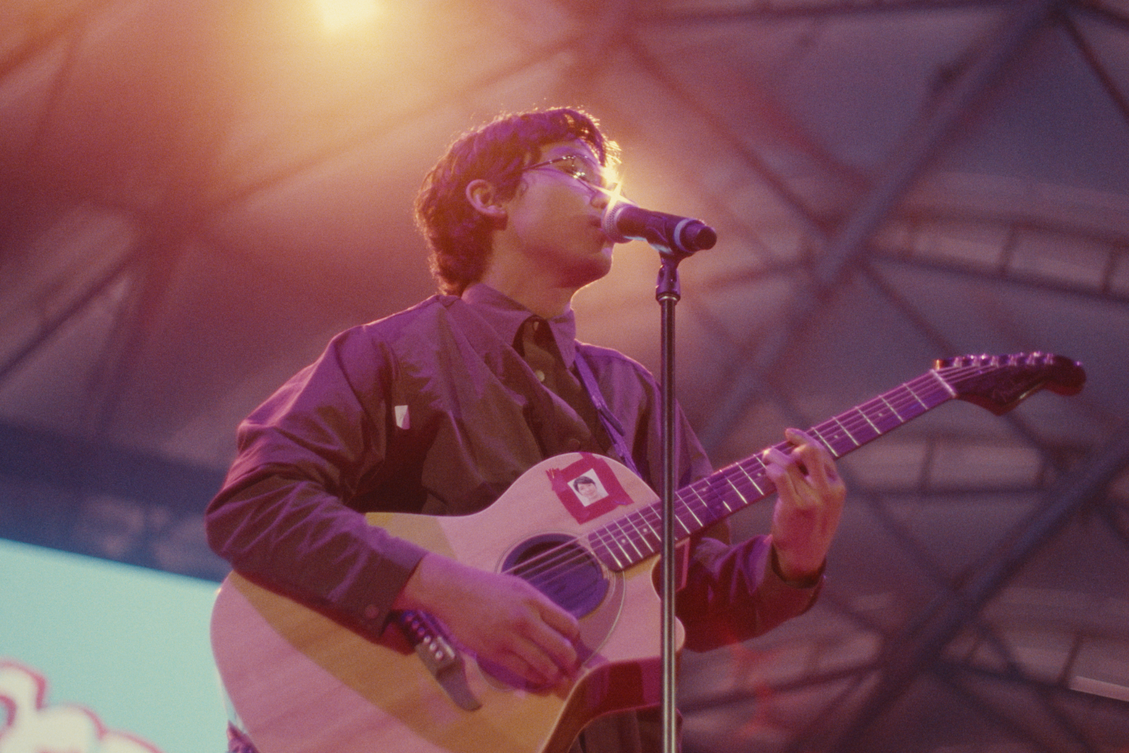 Young man with glasses playing an acoustic guitar on stage, singing into a microphone, under warm stage lighting.