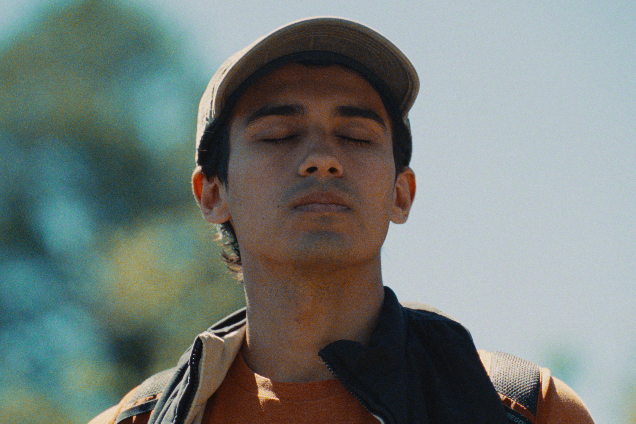 A young man with closed eyes, wearing a cap and a black jacket with a backpack, outdoors on a sunny day.