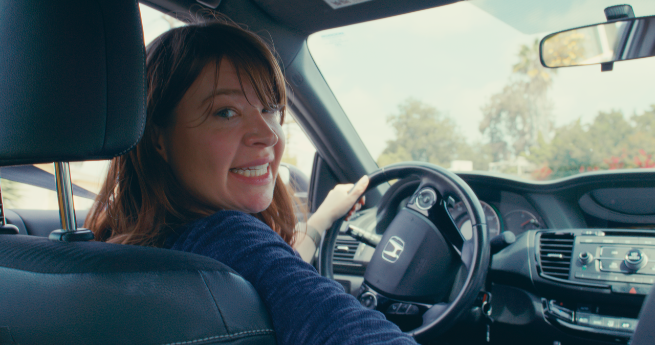 A woman with brown hair smiling and looking back while sitting in the driver's seat of a car, holding the steering wheel with her left hand, with trees and a sunny sky visible through the windshield.