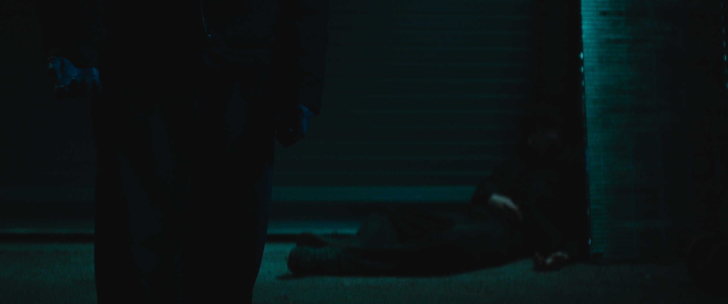 Dark scene with a person kneeling on the floor, another person standing nearby holding a plastic bottle, in a dimly lit room with horizontal blinds.