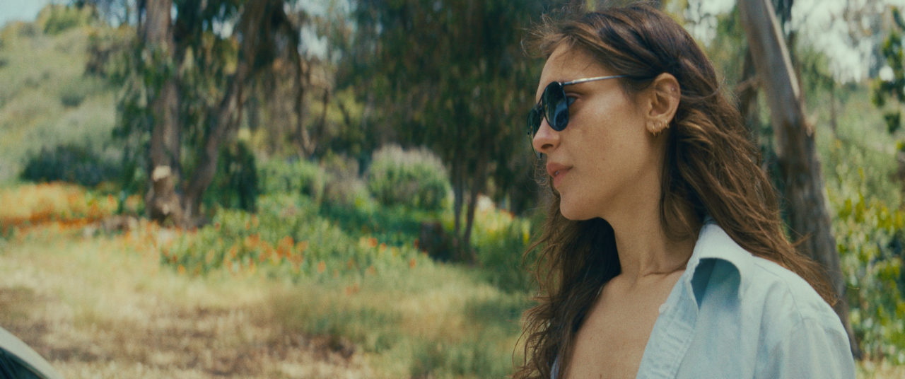 A woman with long curly hair wearing sunglasses and a light blue shirt, standing outdoors in a park or garden with trees and greenery in the background.