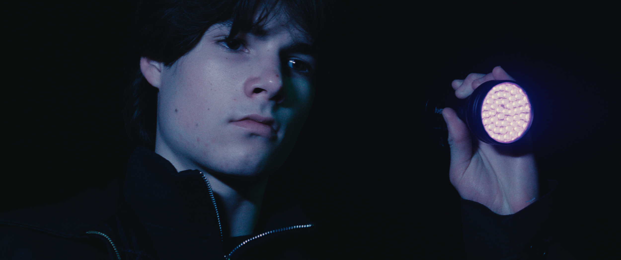 A young man with dark hair and pale skin holding a purple LED flashlight in a dark environment, looking at the camera with a serious expression.