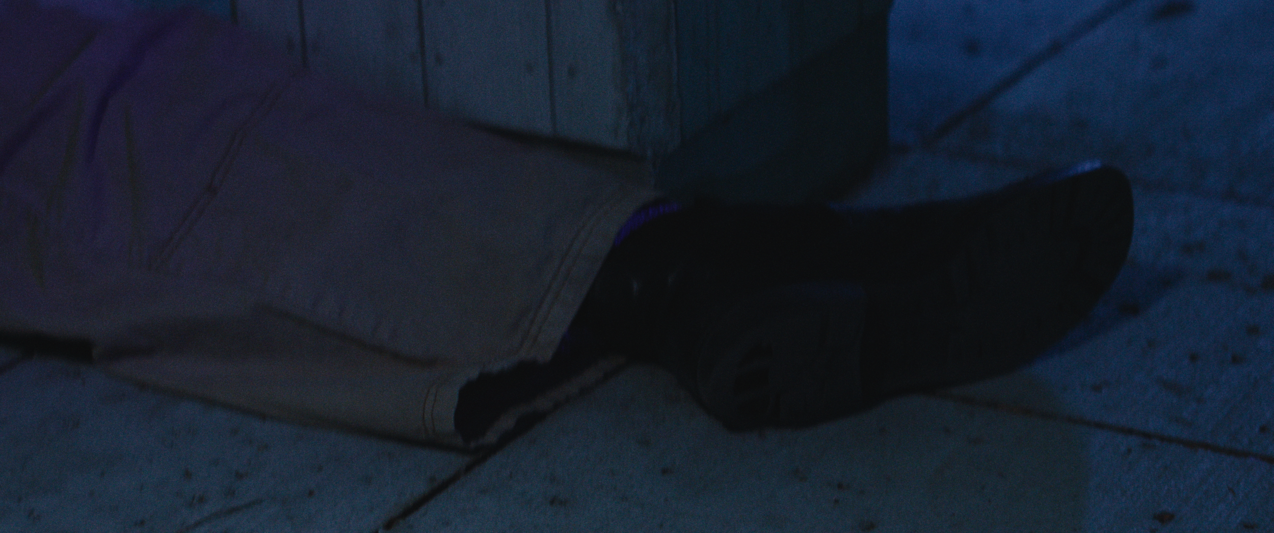Close-up of a person's lower leg and shoe, taken in a dark setting, with gray pants and black footwear visible on concrete tiles.