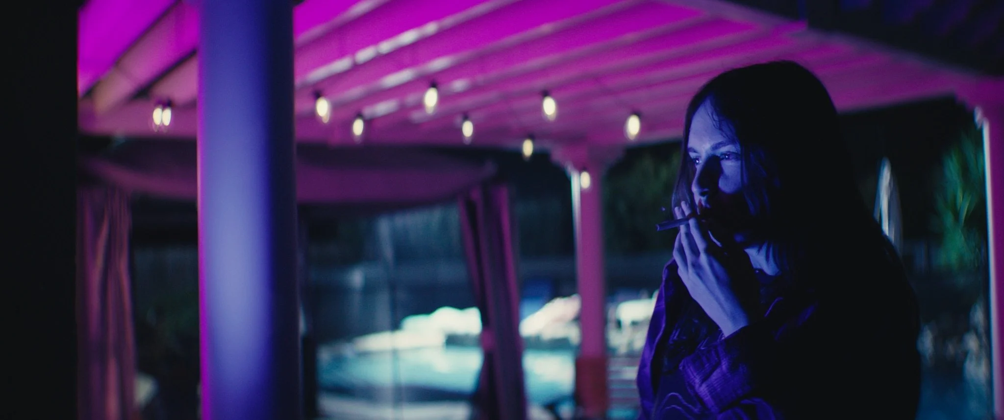 A woman with long dark hair is smoking a cigarette in a dimly lit area with purple and blue lighting, under a wooden roof with string lights.