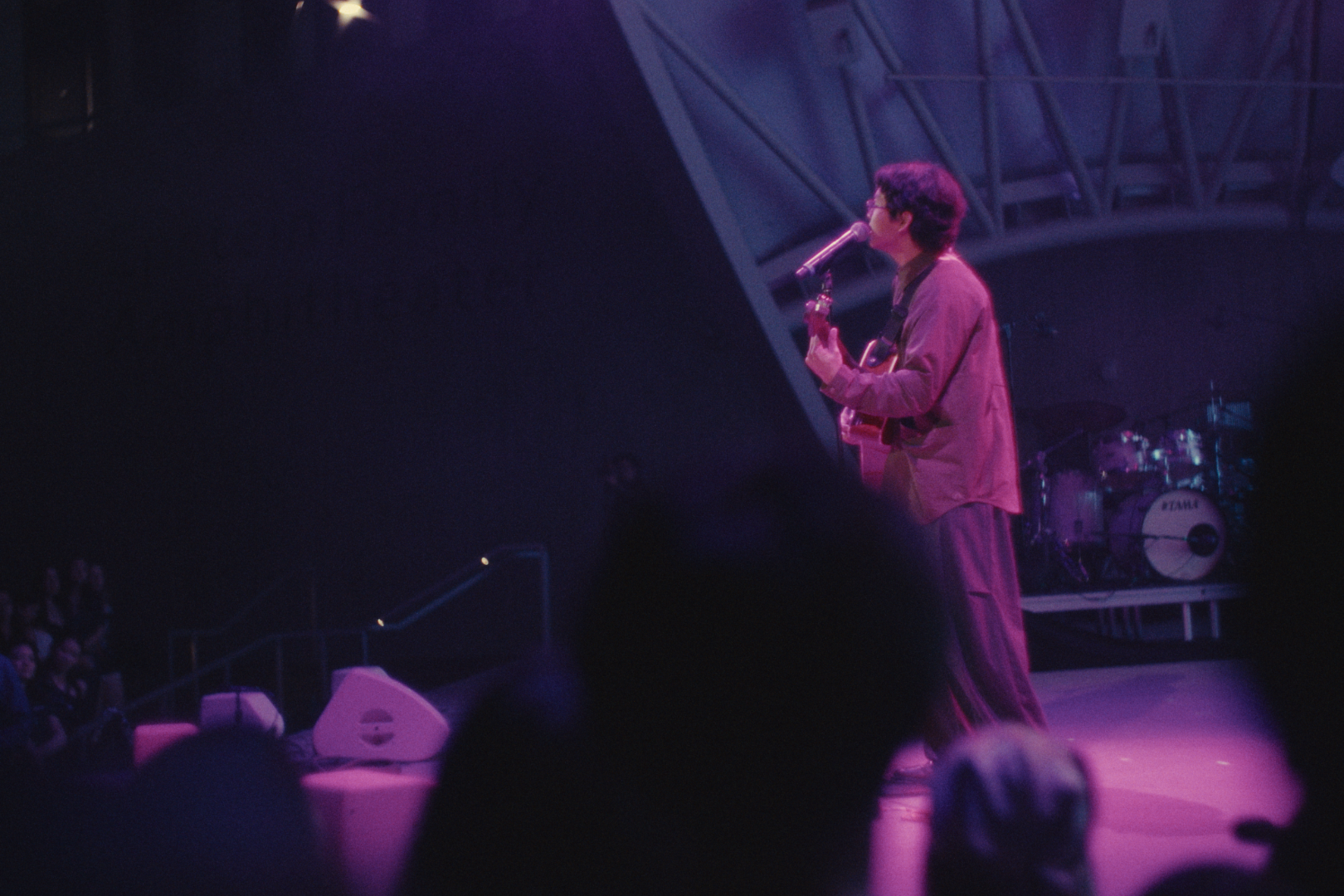 A man performing on stage with a guitar, singing into a microphone, with drums in the background, illuminated by pink and purple stage lights.