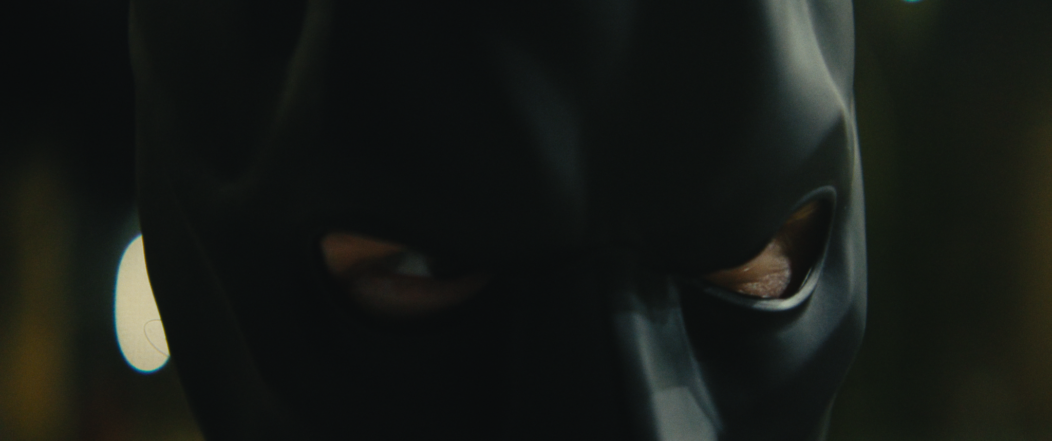 Close-up of a person wearing a black mask with eye holes, looking downward.