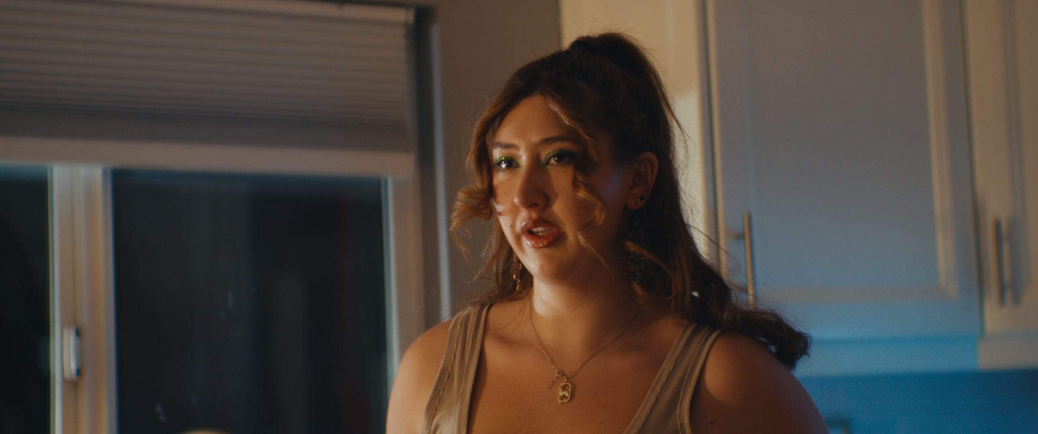 A woman with shoulder-length brown hair and green eyeshadow wearing a beige tank top, jewelry, and a necklace, standing in a room with her lips parted, looking to the side.