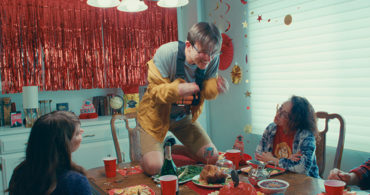 A man stands on a dining table, smiling and celebrating with friends. The table is decorated with a roasted turkey, red cups, food, and confetti, indicating a festive gathering or party.