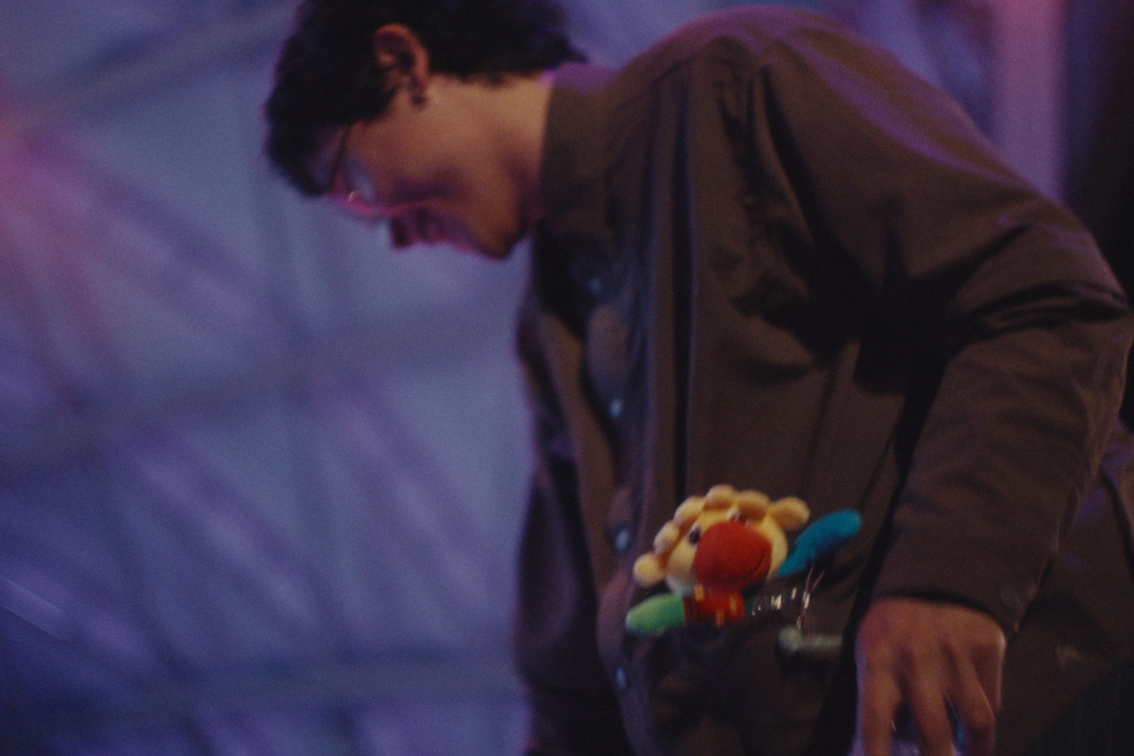 A person wearing glasses, a brown jacket with a plush toy of a yellow lion with an orange nose in the jacket's pocket, and a blue plush toy in the pocket, bent over with their mouth open against a dark purple background.