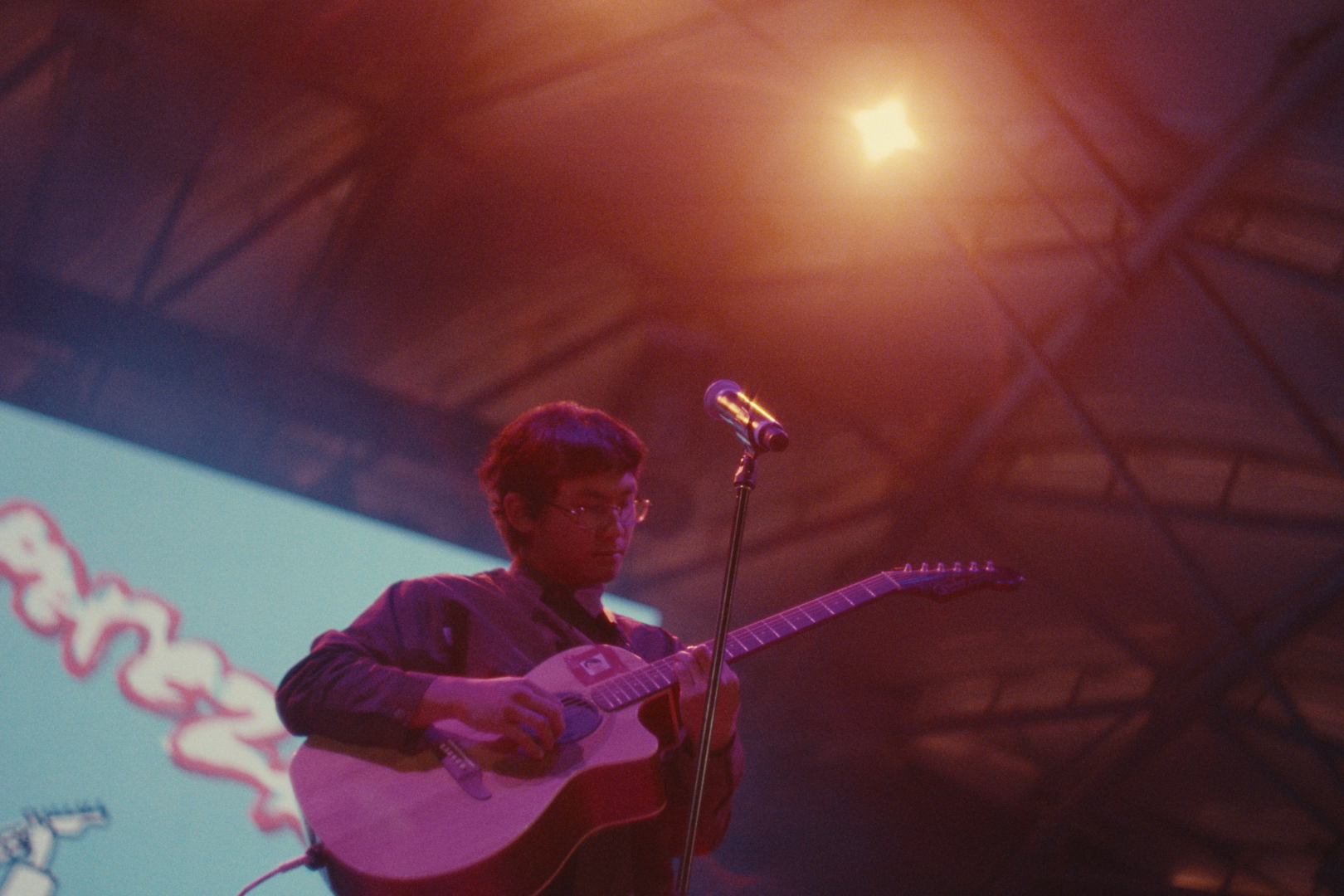 A person wearing glasses playing an acoustic guitar on stage under warm lighting.