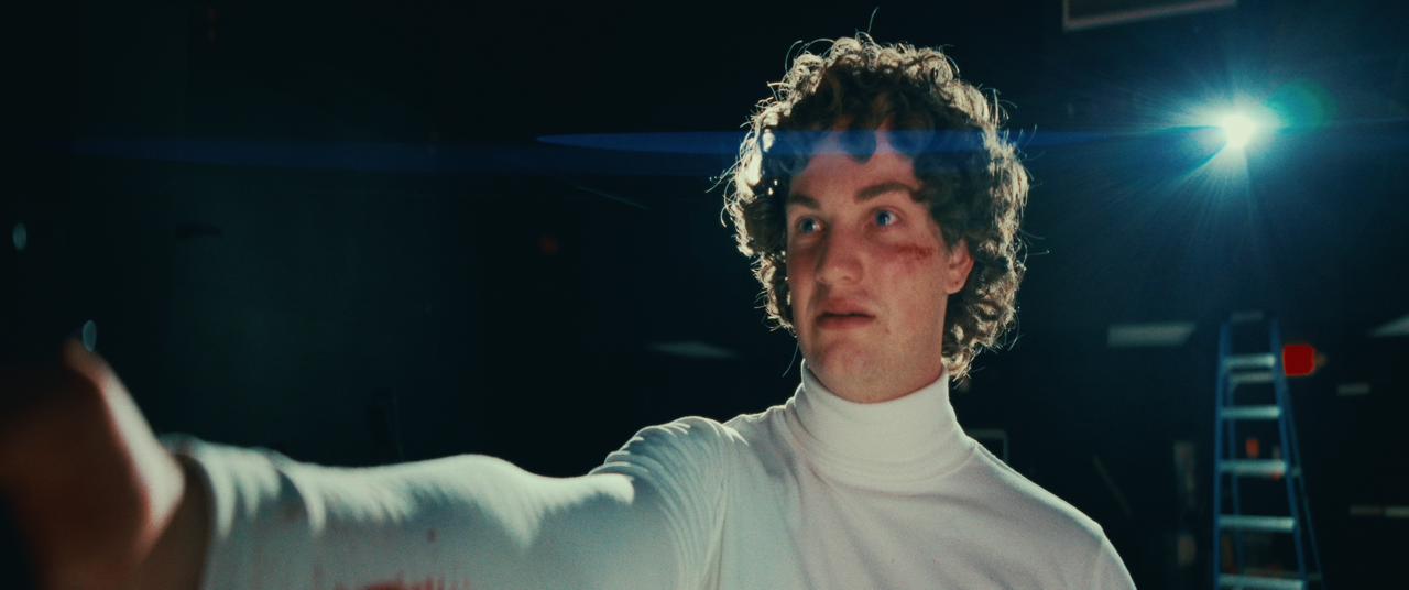 A young man with curly hair and a white turtleneck, looking shocked or surprised, standing in a dimly lit room with bright lights in the background.