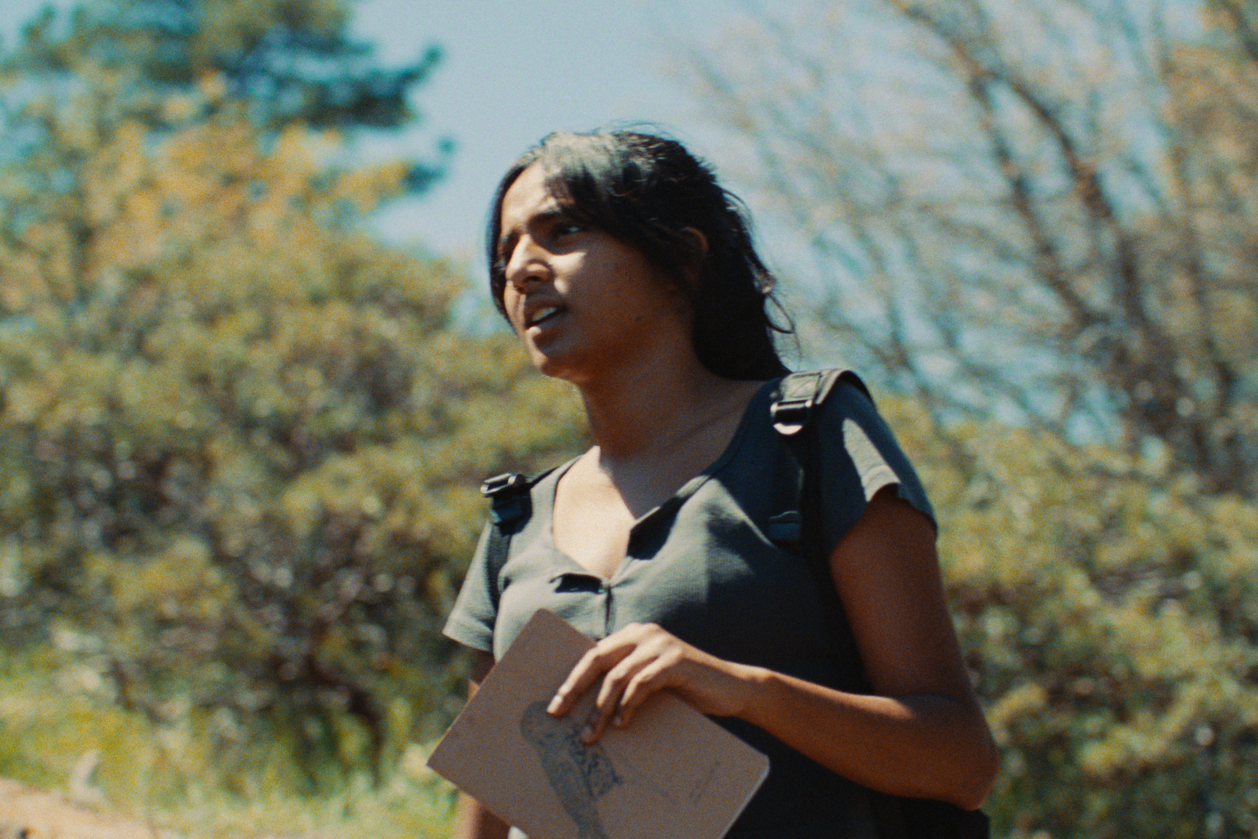 A woman with long black hair, wearing a striped shirt, holding a sketchbook with a drawing, outdoors surrounded by trees.
