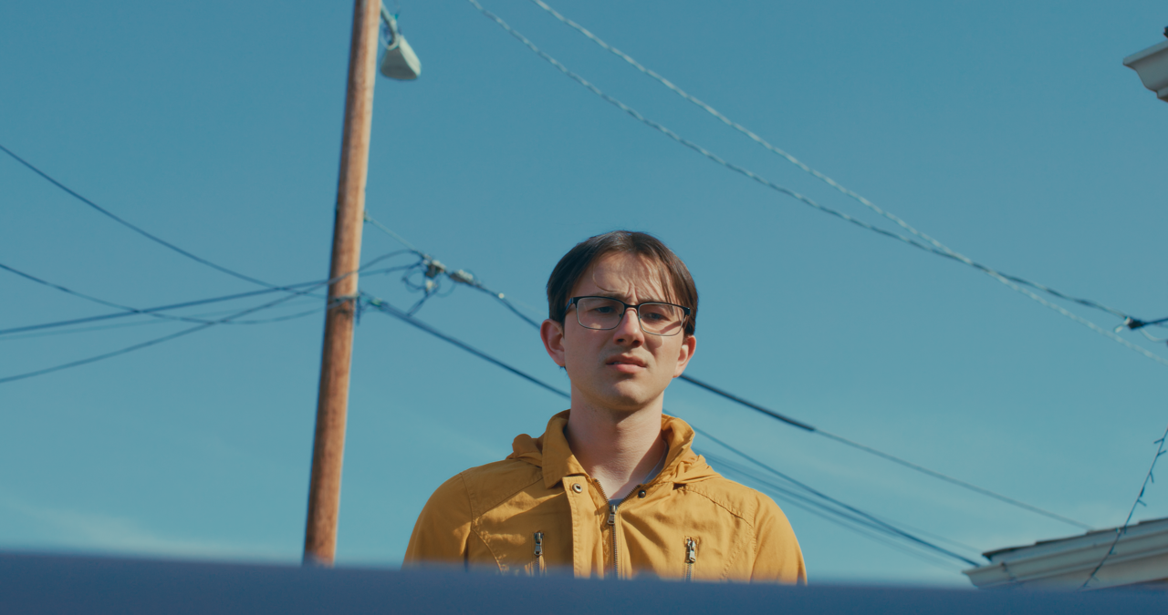 A young man with glasses wearing a yellow jacket standing outdoors under power lines against a clear blue sky.