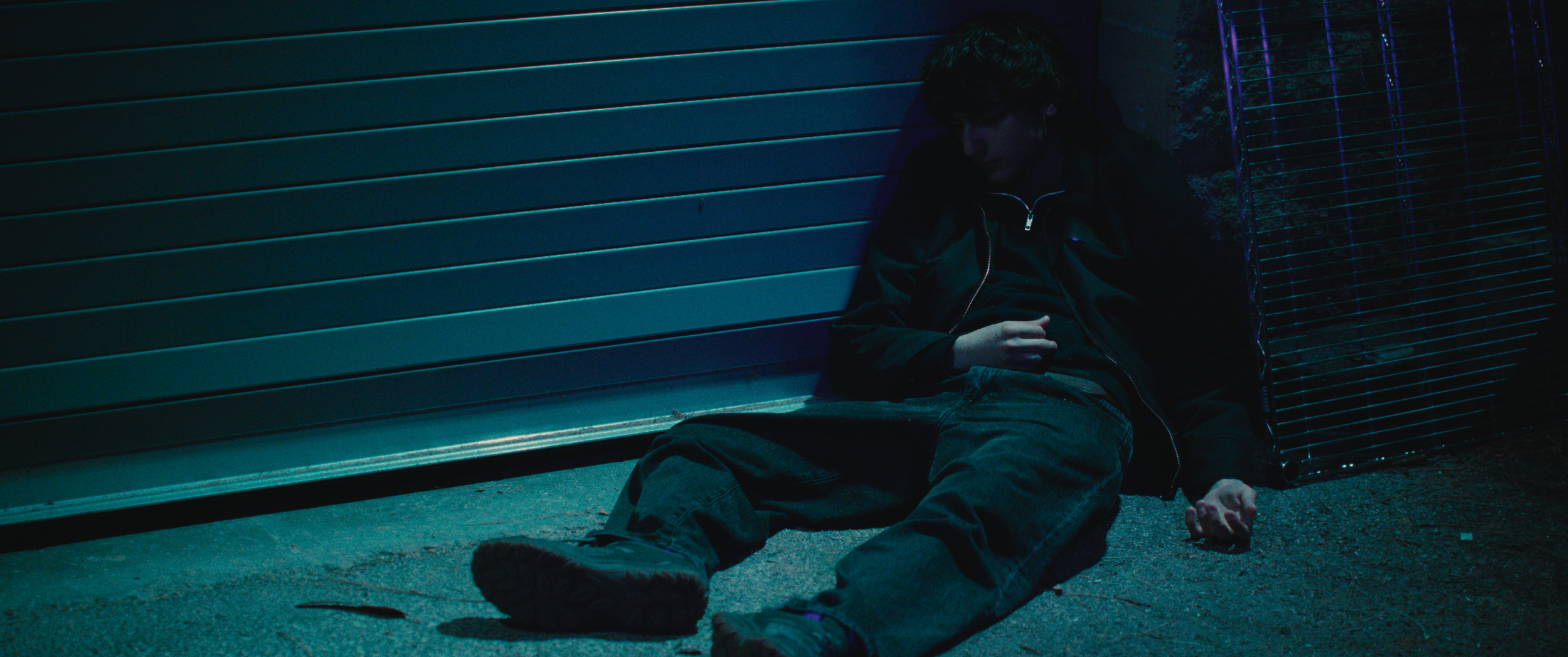 A young man sitting on the ground against a building with a metal garage door at night. He is wearing a dark jacket and jeans, with his head bowed and eyes closed, appearing to be exhausted or in distress.