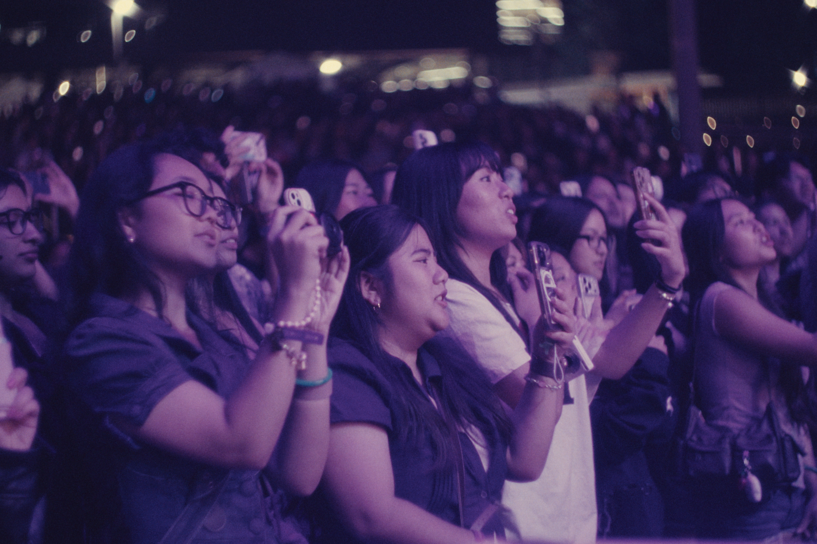 Crowd of women at an event, some taking photos or videos with their smartphones, under purple lighting.